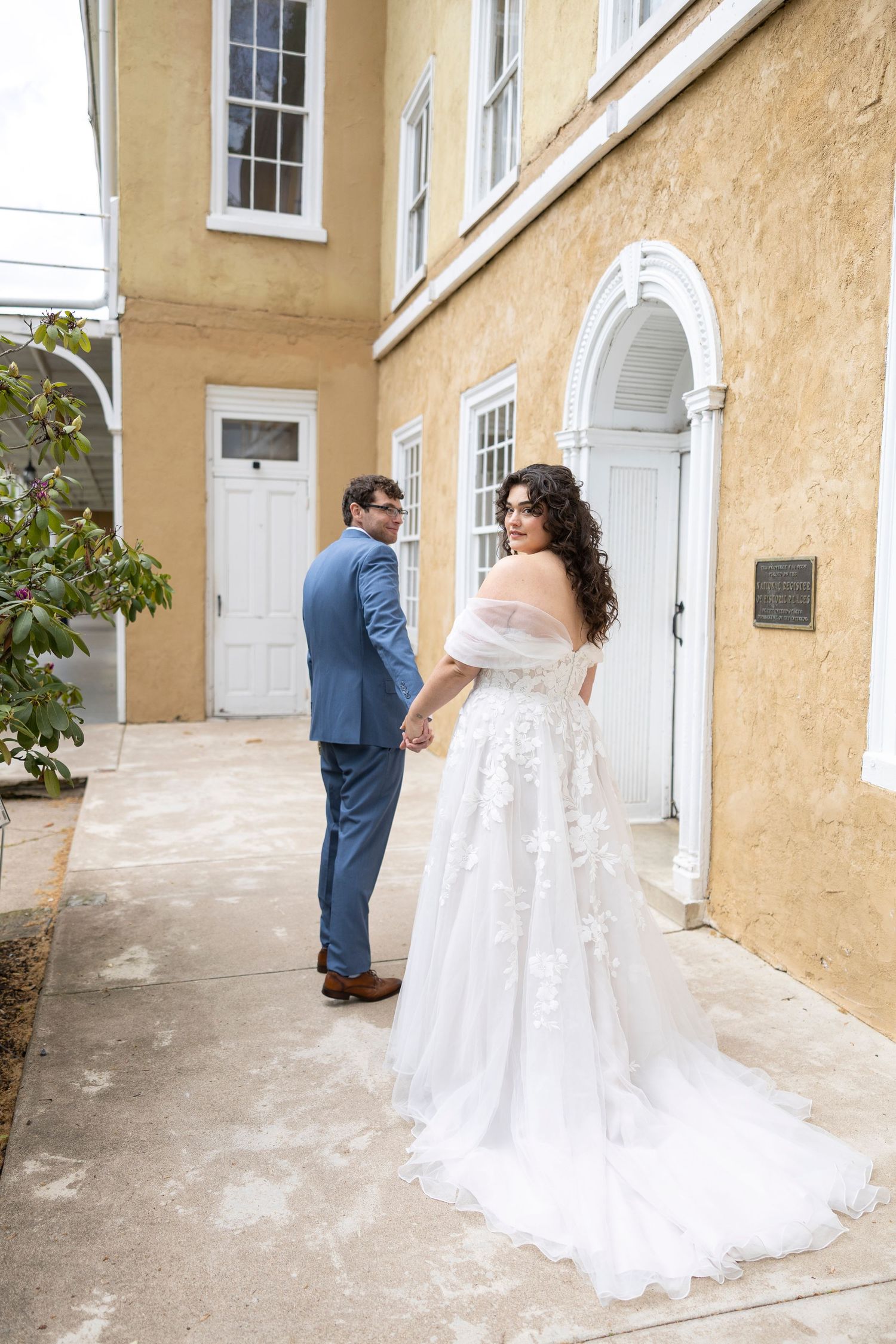 Bride in off-shoulder floral gown and groom in blue suit hold hands outside a yellow historic building.