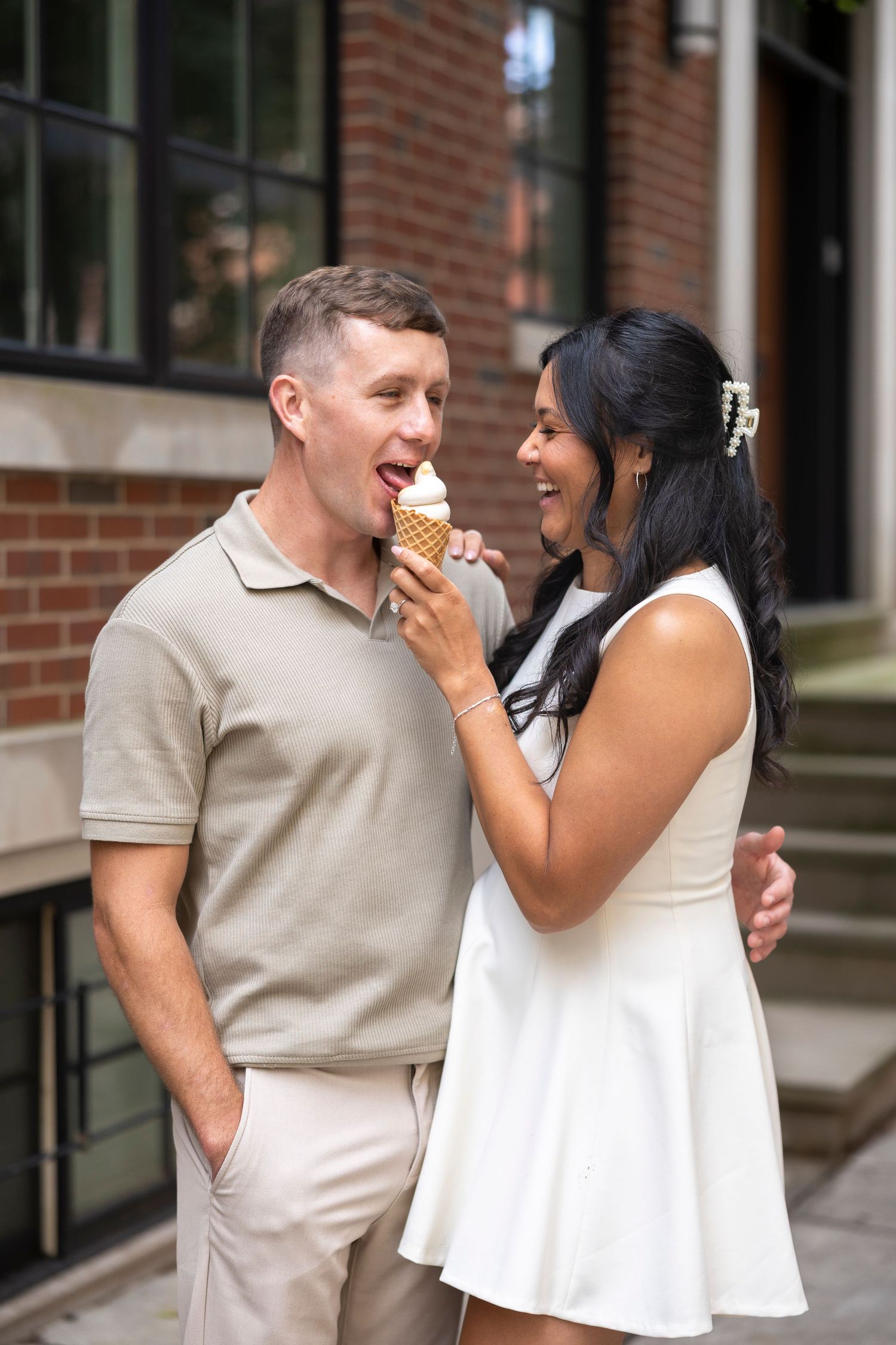 A couple shares a lighthearted moment while eating ice cream outside a brick building.