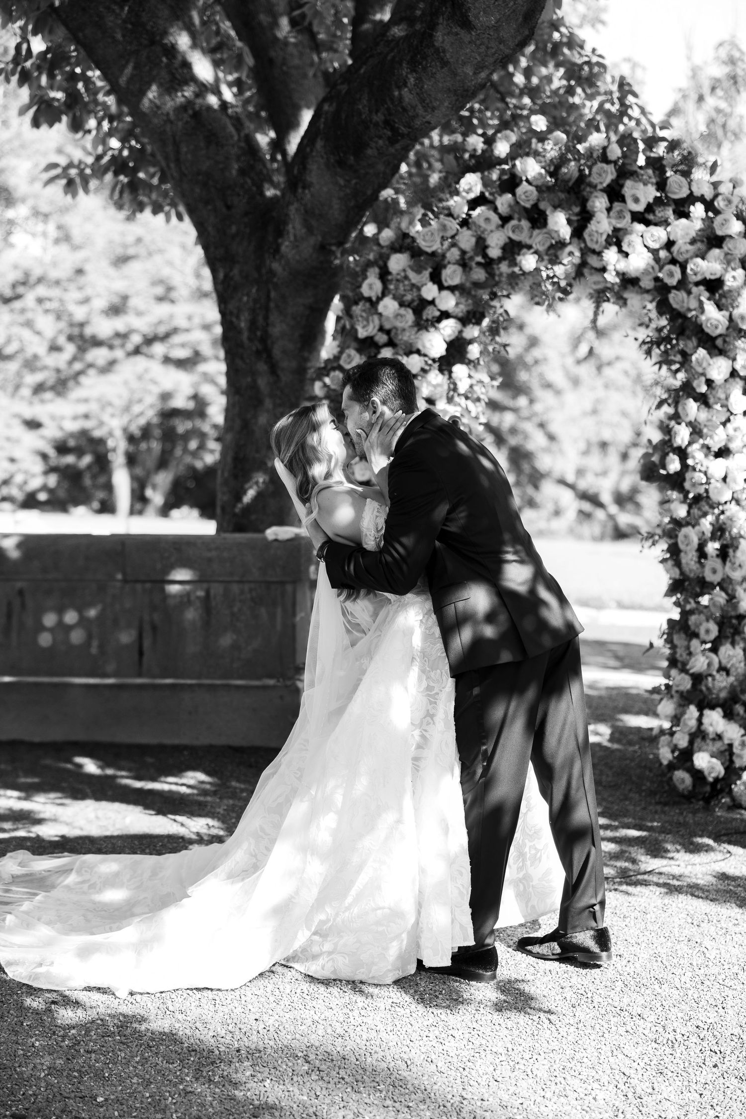 Black and white photo of a wedding ceremony under a floral arch in natural light.