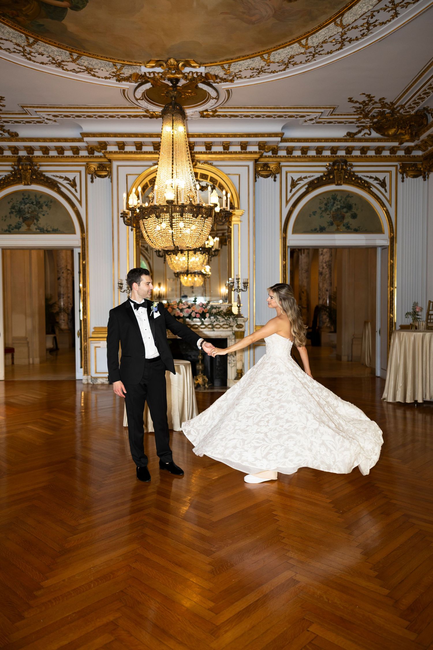 Couple dances in an ornate ballroom with crystal chandeliers and herringbone floors.
