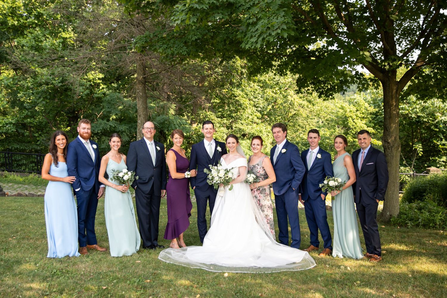 Wedding party poses outdoors in coordinated navy and pastel dresses against lush greenery.