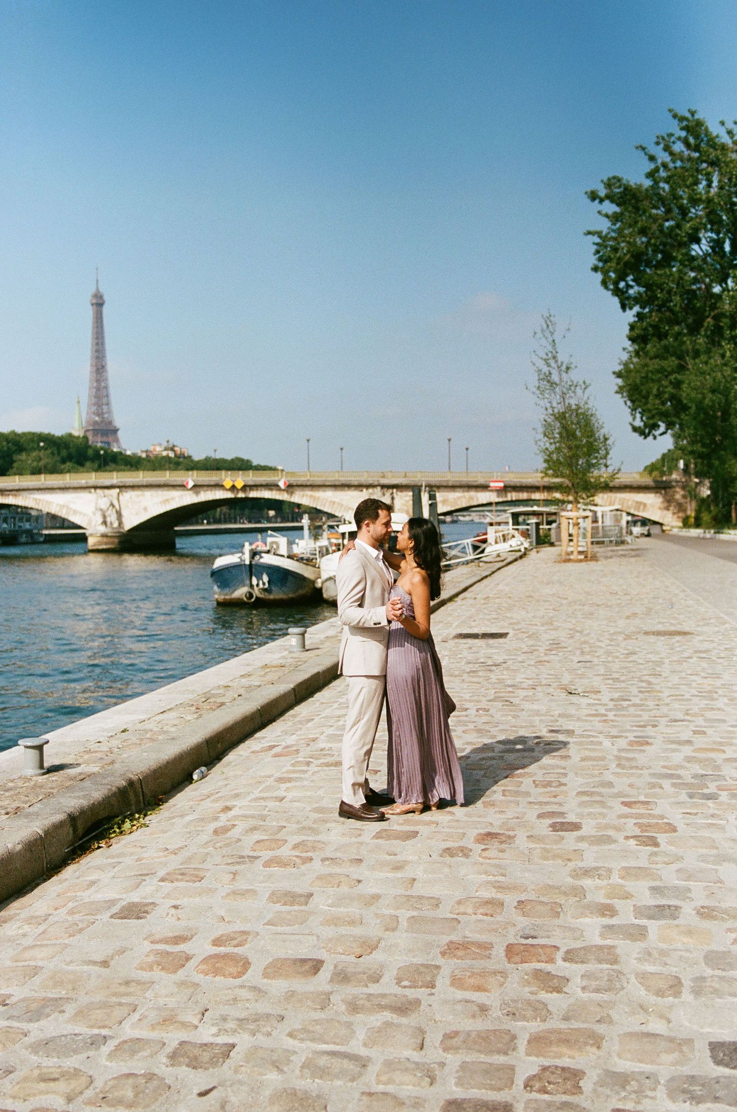 Romantic couple photos along Seine River with Eiffel Tower in background on cobblestone path.