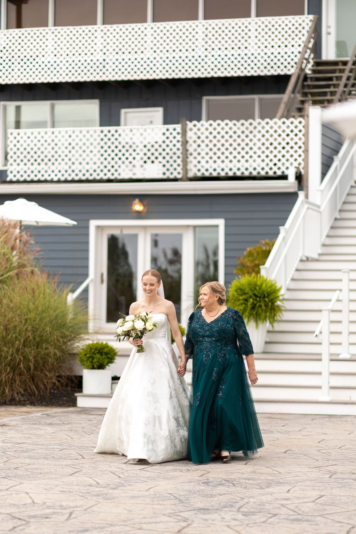 A bride and companion walk together on a stone path in front of a coastal venue with white stairs.