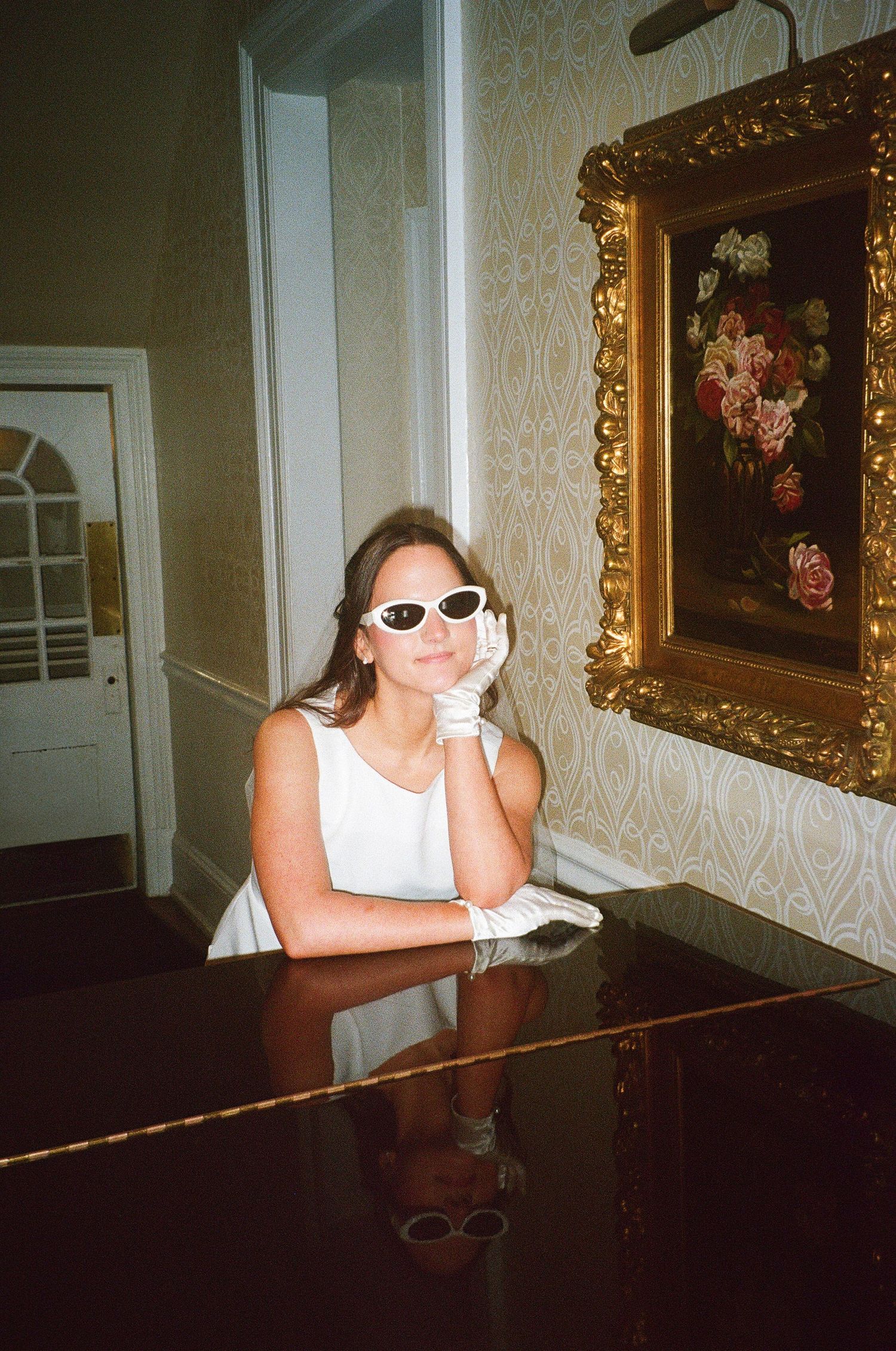 Person in white dress and sunglasses posing at grand piano in elegant room with ornate decor.