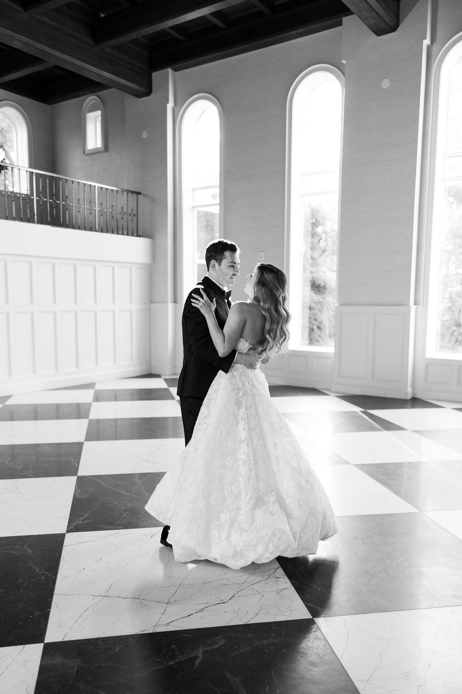 A couple in formal wedding attire dances in an elegant room with a black and white checkered floor and bright windows.
