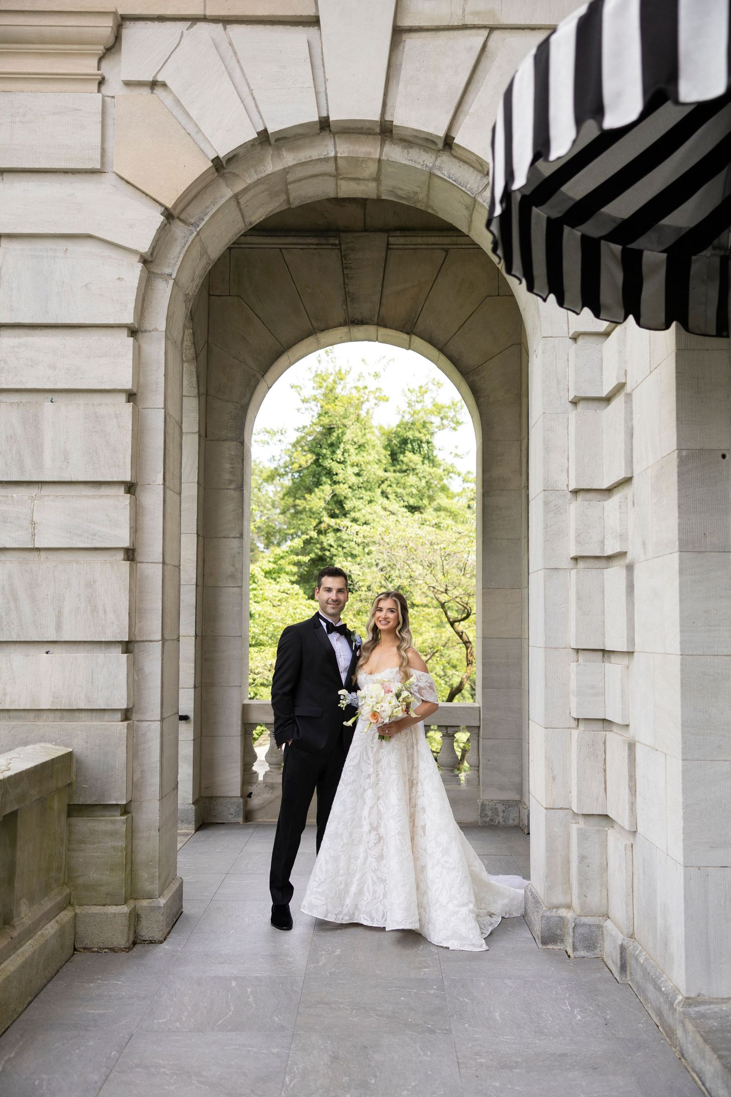 Wedding couple walks through stone archway corridor with classical architecture.