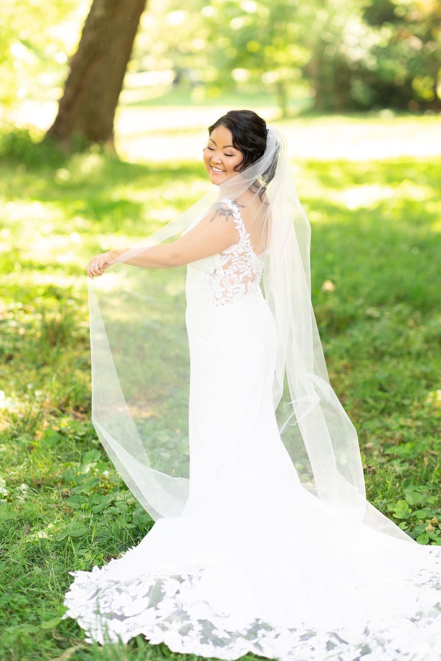 A bride in a flowing lace gown twirls her veil in a sun-dappled garden setting.