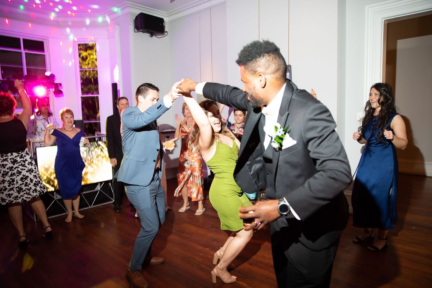 Wedding guests dancing and celebrating at a lively indoor reception with colorful lighting.