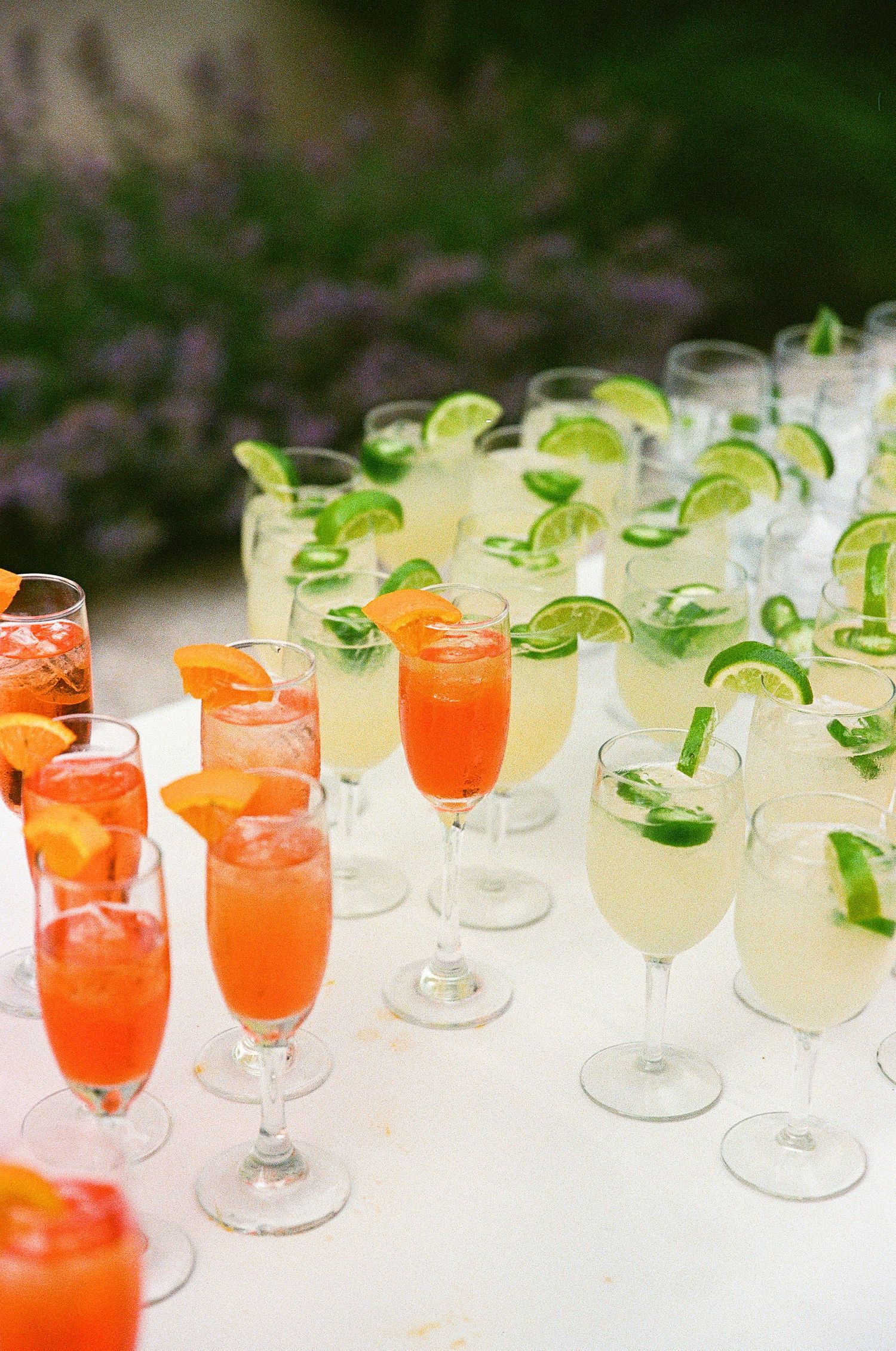 Elegant display of orange and lime garnished cocktails in champagne flutes arranged on a white table at an outdoor event.