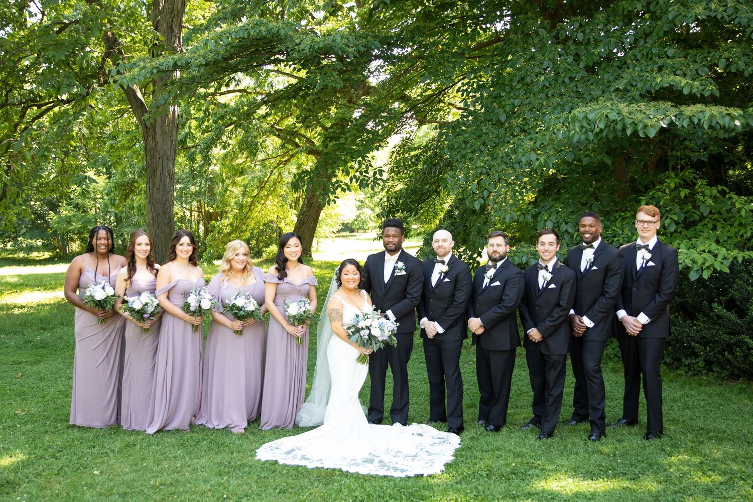 A wedding party in purple dresses and black tuxedos poses together under green trees.
