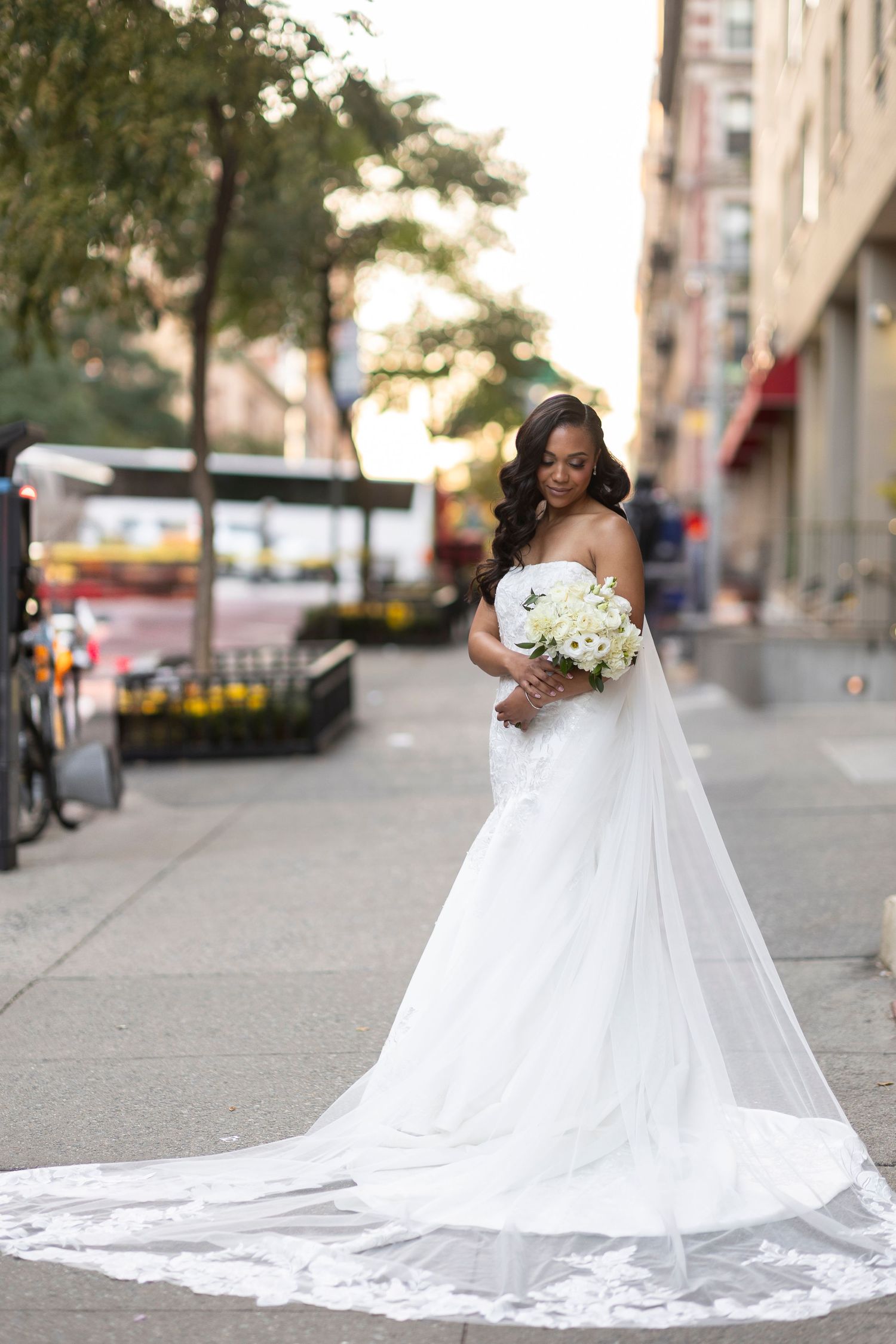 Bride in flowing white gown with cathedral train holds bouquet on city sidewalk at sunset.