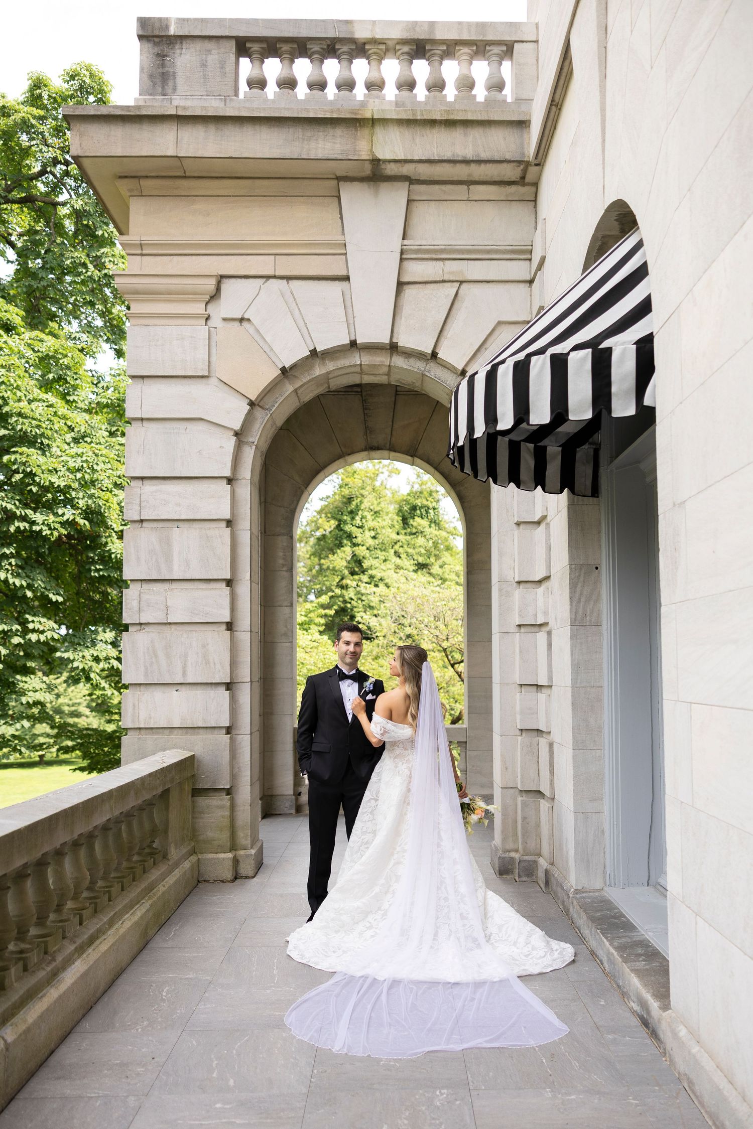 Elegant wedding couple poses beneath a stone archway with striped awnings in the background.