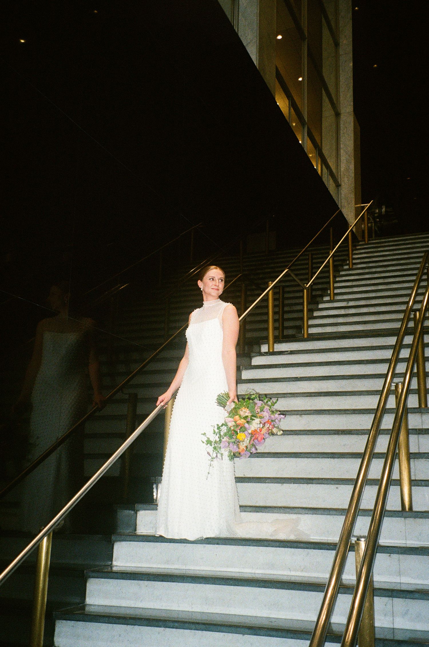 Bride in white gown holding colorful bouquet stands on marble stairs with gold railings at elegant venue.