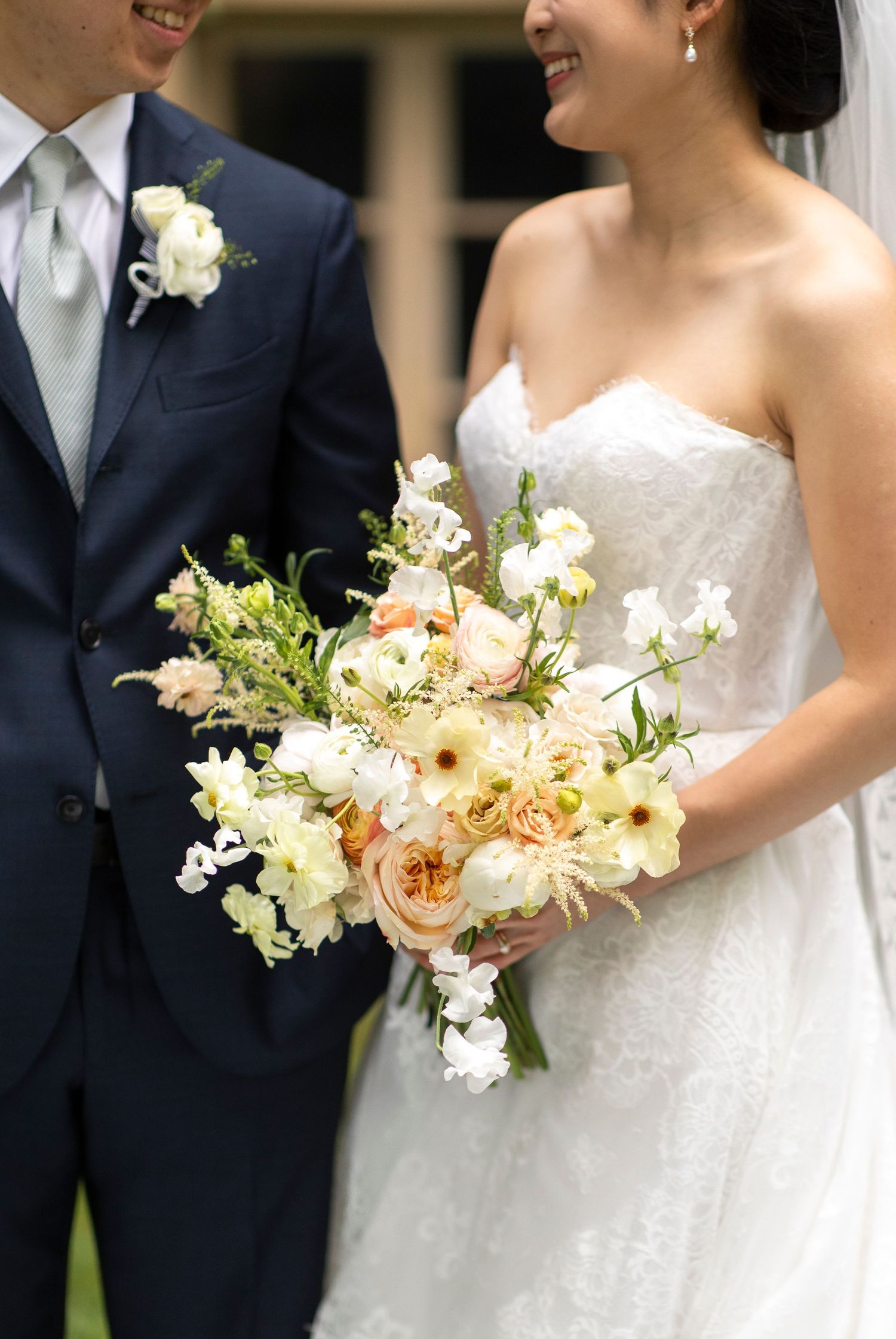 Bride in strapless lace gown holds lush peach and white bouquet beside groom in navy suit with boutonniere.