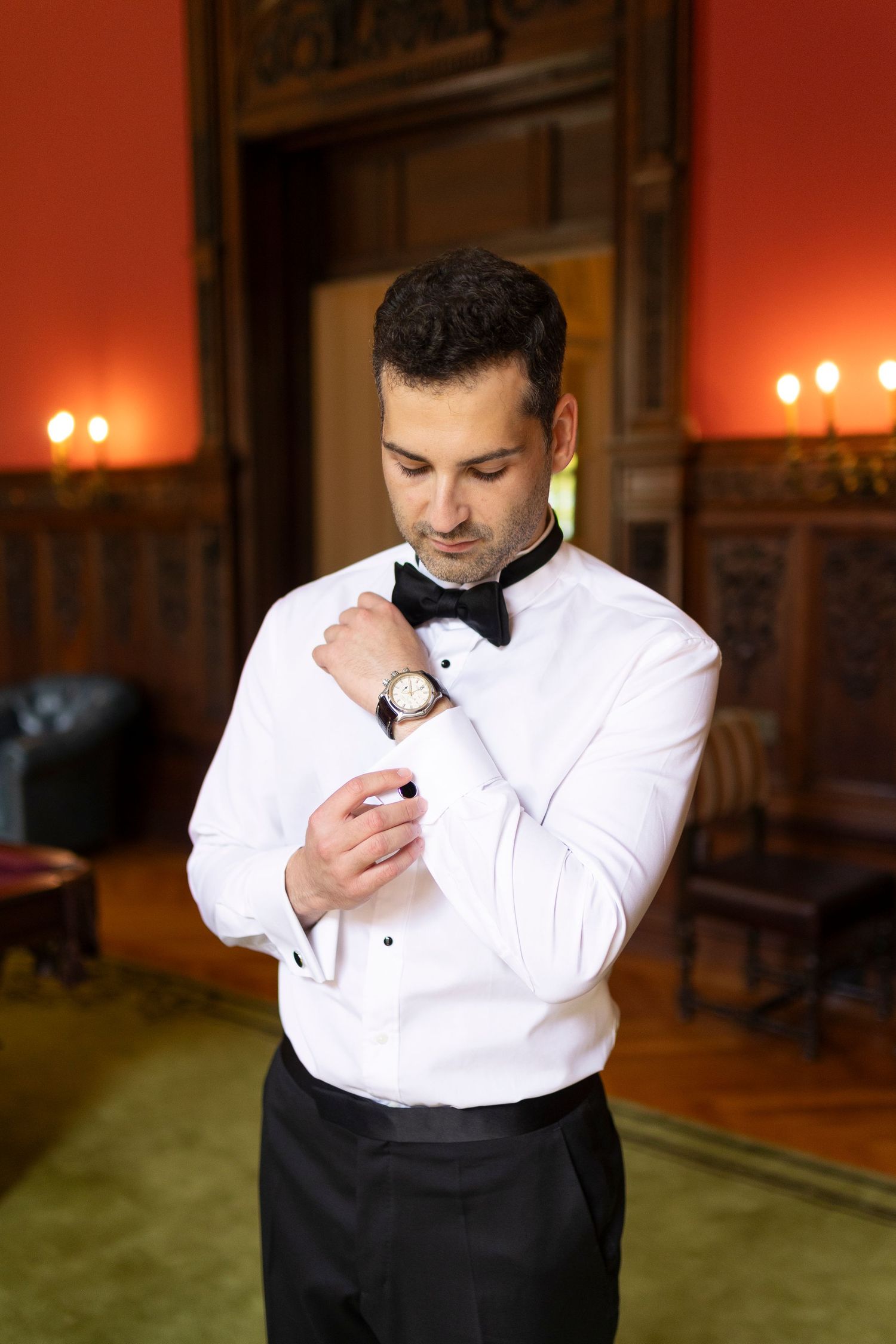 A dapper groom in a white tuxedo shirt and black bow tie adjusts his cufflinks in an elegant wood-paneled room.