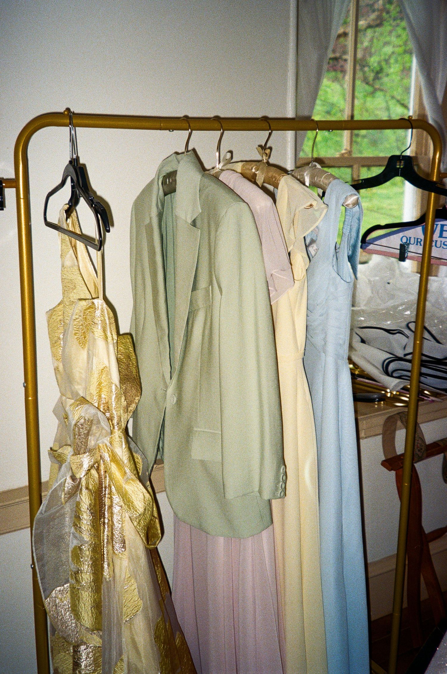 Pastel formal dresses and a green blazer hanging on a gold clothing rack near a window.