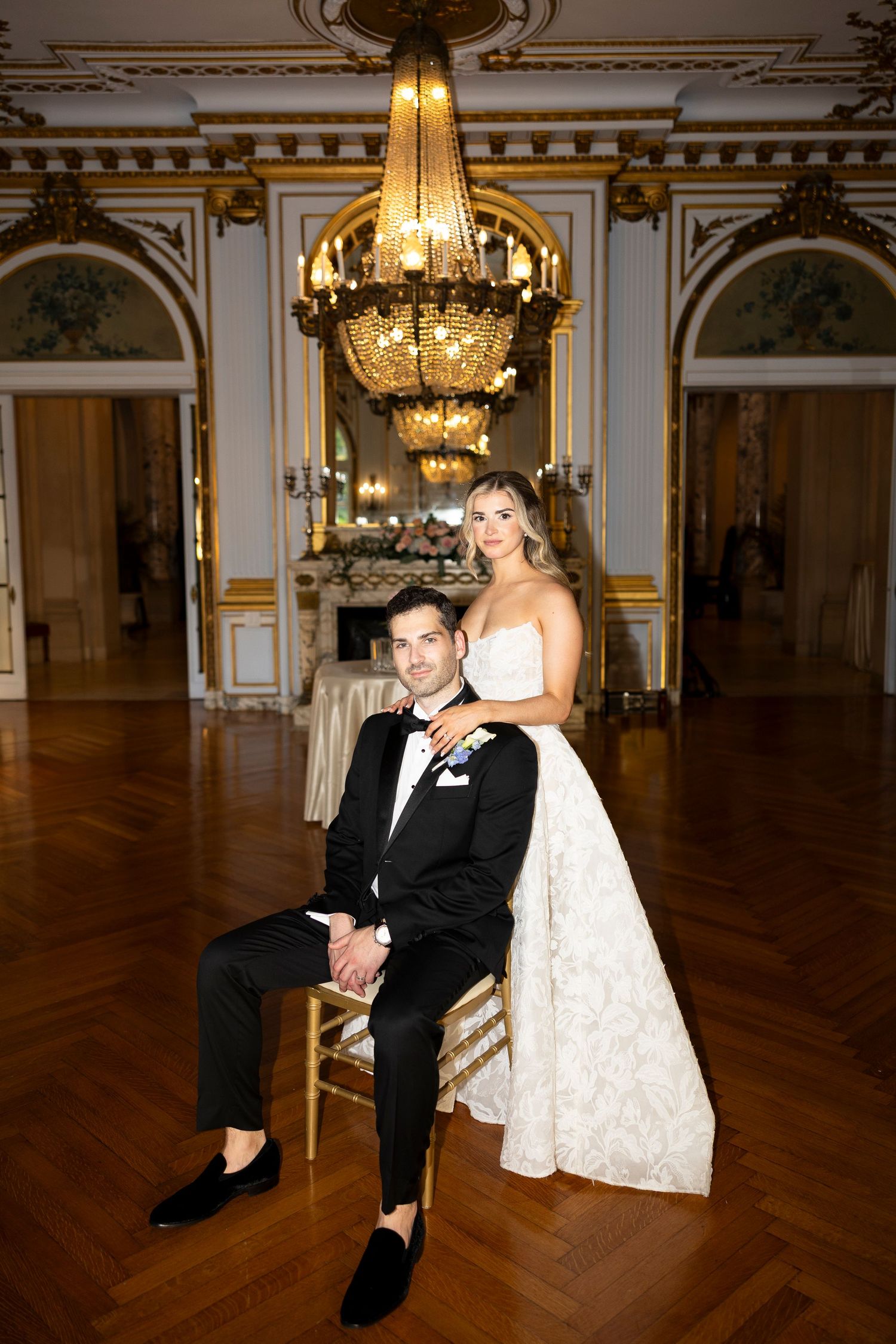 Elegant bride and groom posing in ornate gold ballroom, groom seated in chair with bride standing beside him.