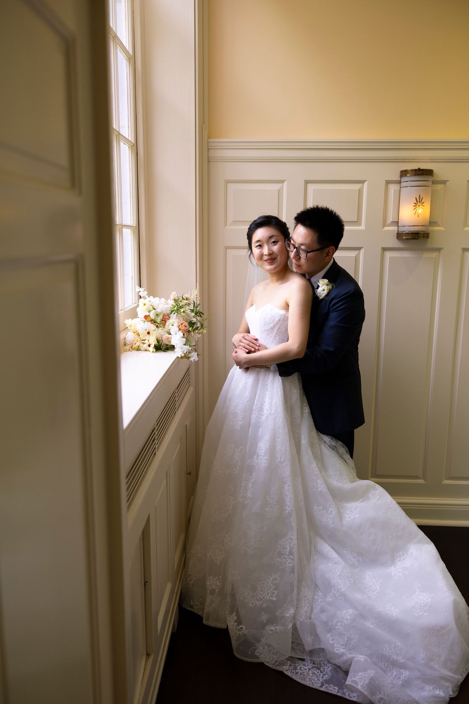A bride and groom share a tender embrace by a window, she in a white lace gown, he in a navy suit, with a floral bouquet nearby.