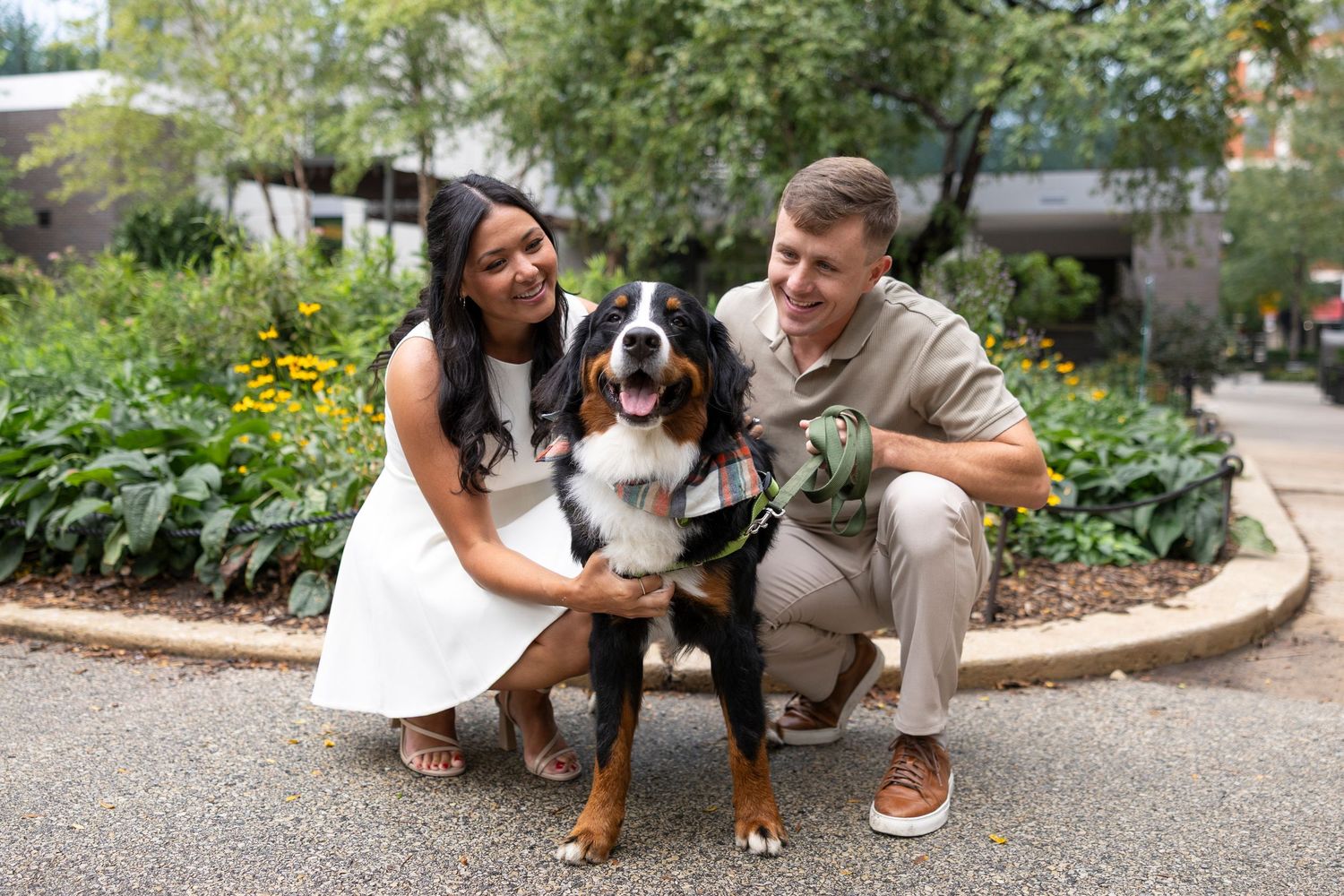 A couple kneels beside their Bernese Mountain Dog on a gravel path.