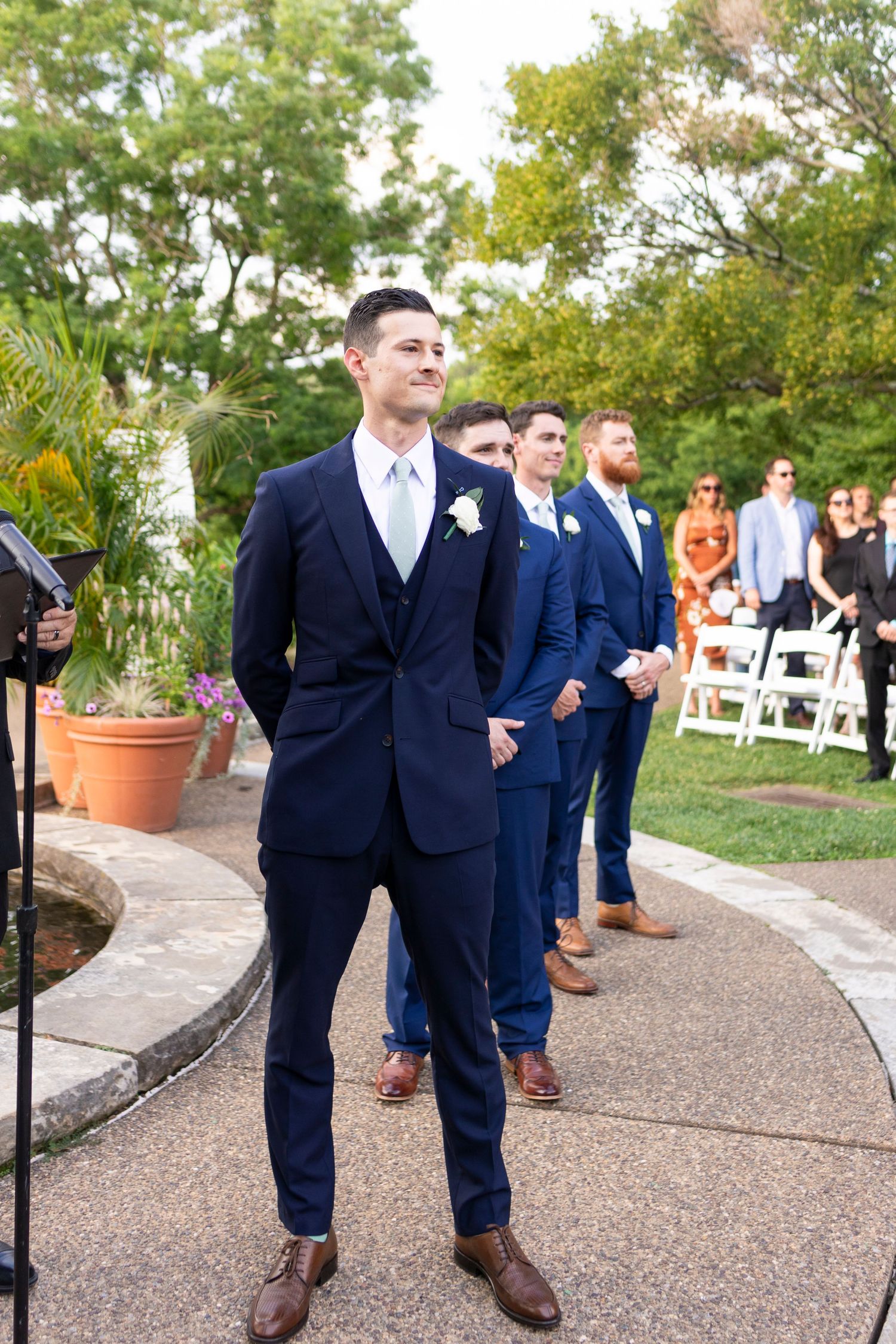 Groomsmen in navy suits and brown shoes line up on a curved garden pathway.
