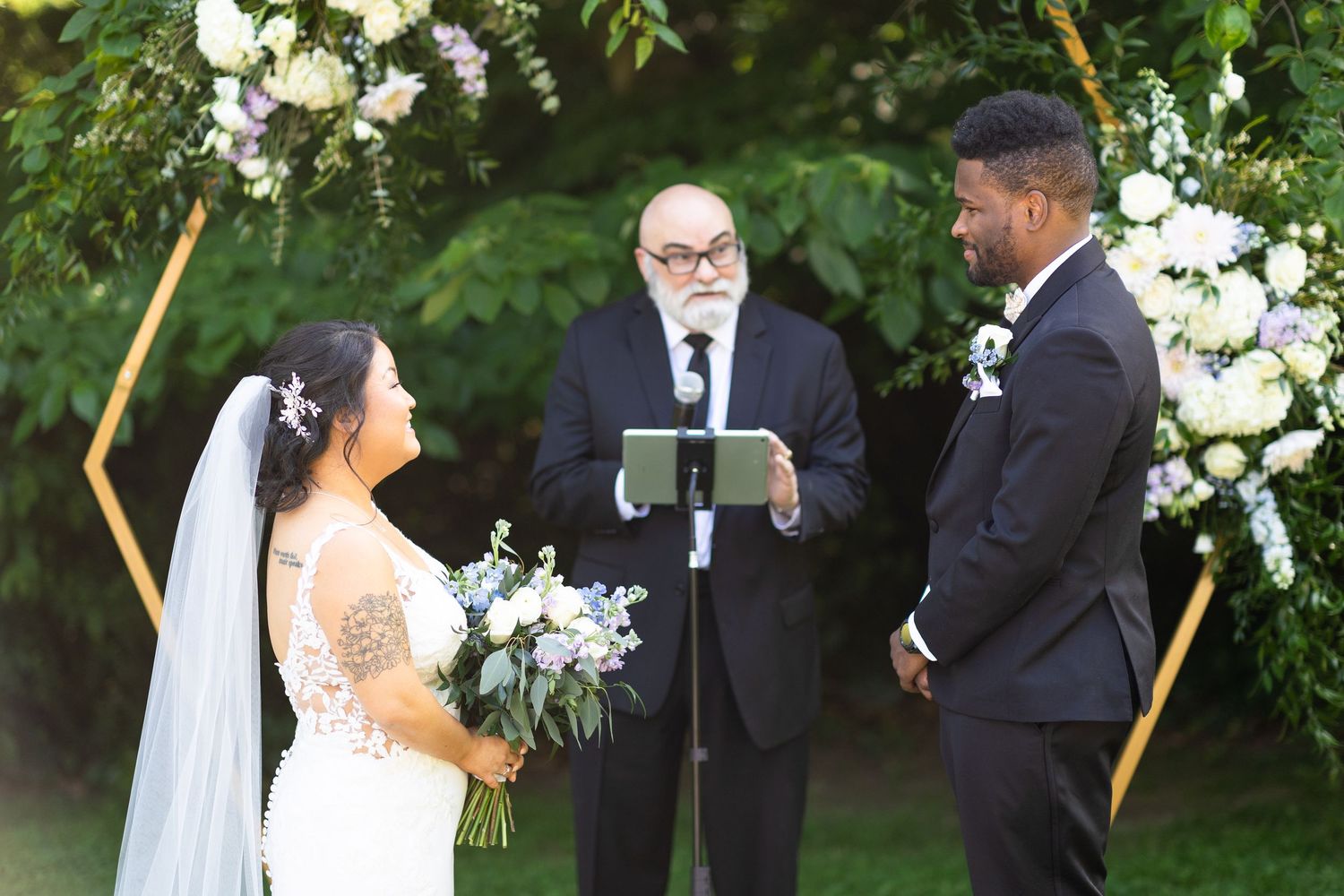 Outdoor wedding ceremony with white floral arch and bridal party on green lawn.