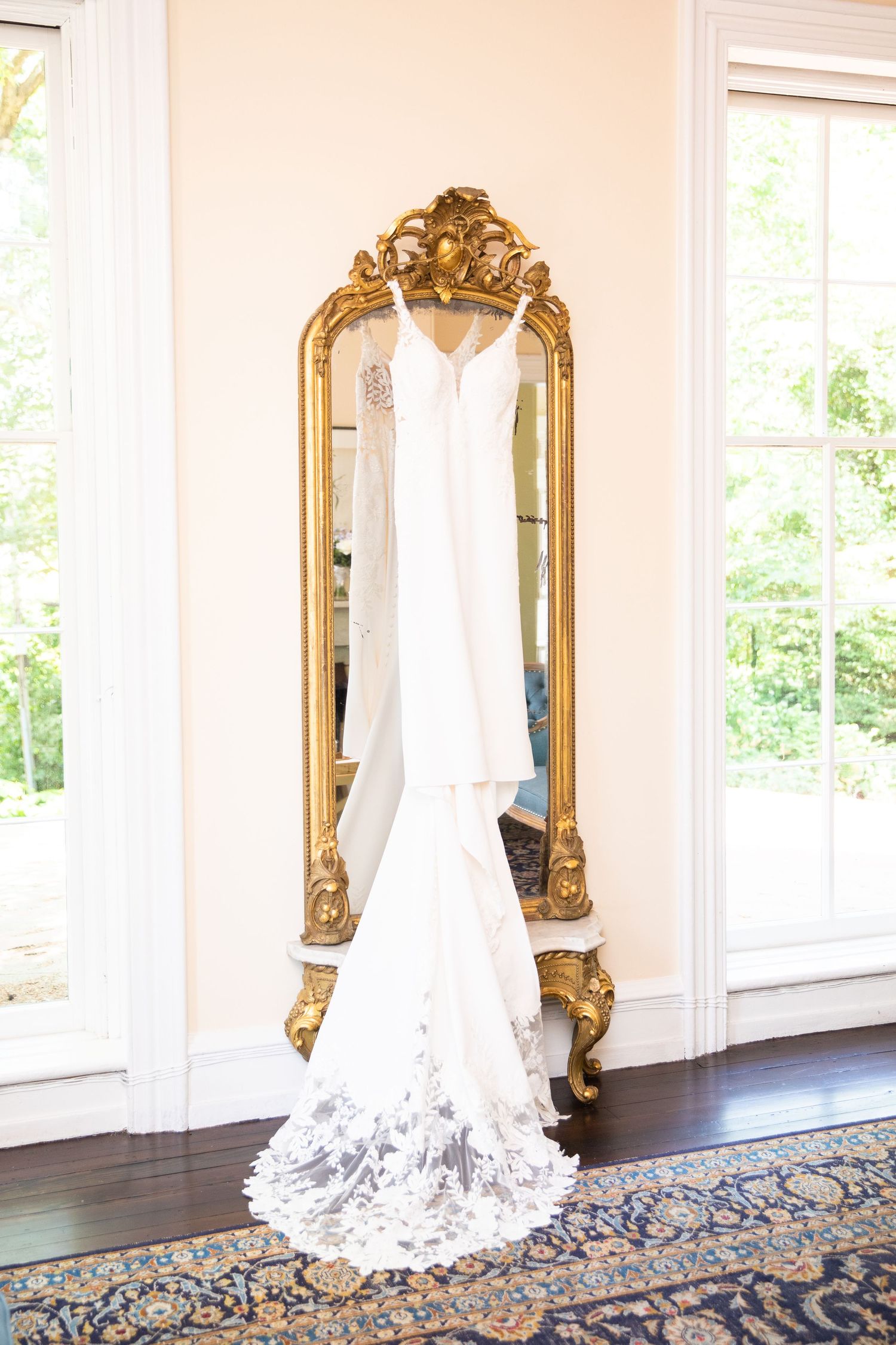 An elegant white wedding dress displayed against ornate gold mirrors in a bright room with hardwood floors.