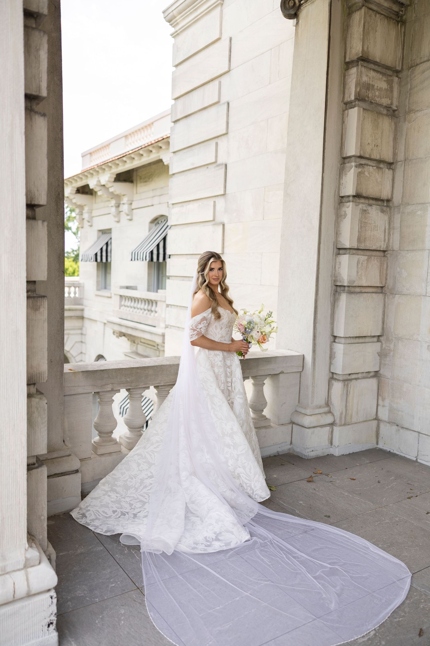 Bride in flowing lace wedding gown with cathedral train posing on classical stone veranda.