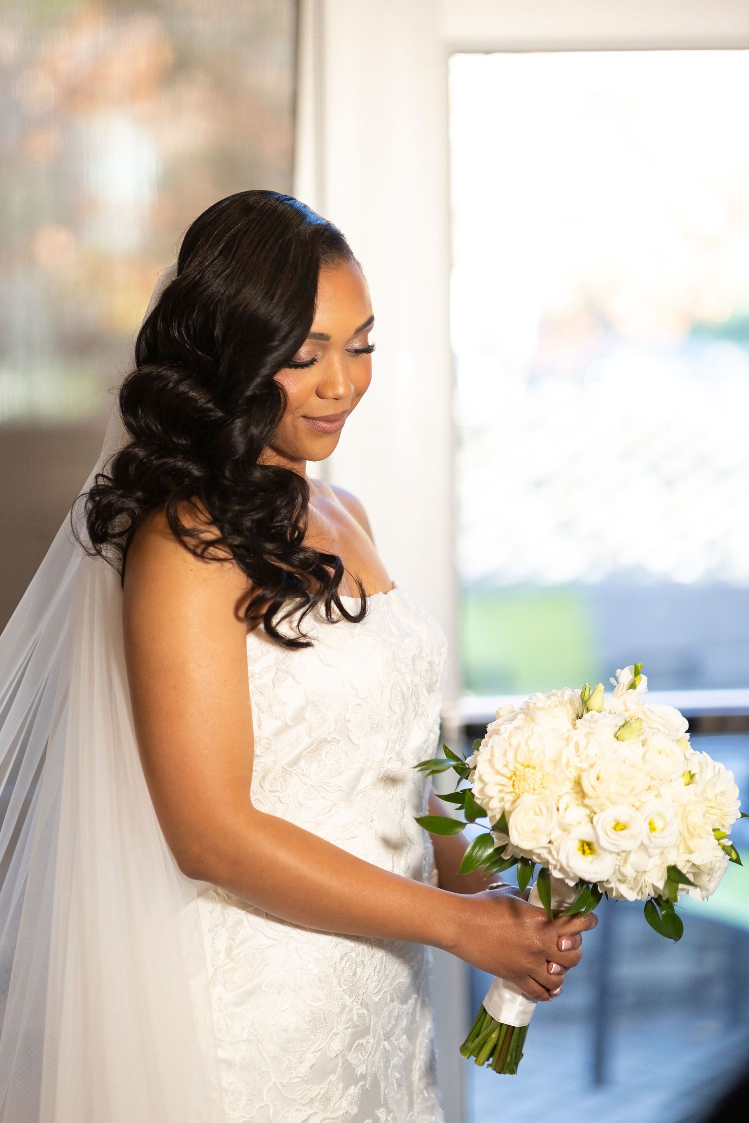 Bride in a white wedding dress holding a bouquet of white flowers while posing near a window with natural light.
