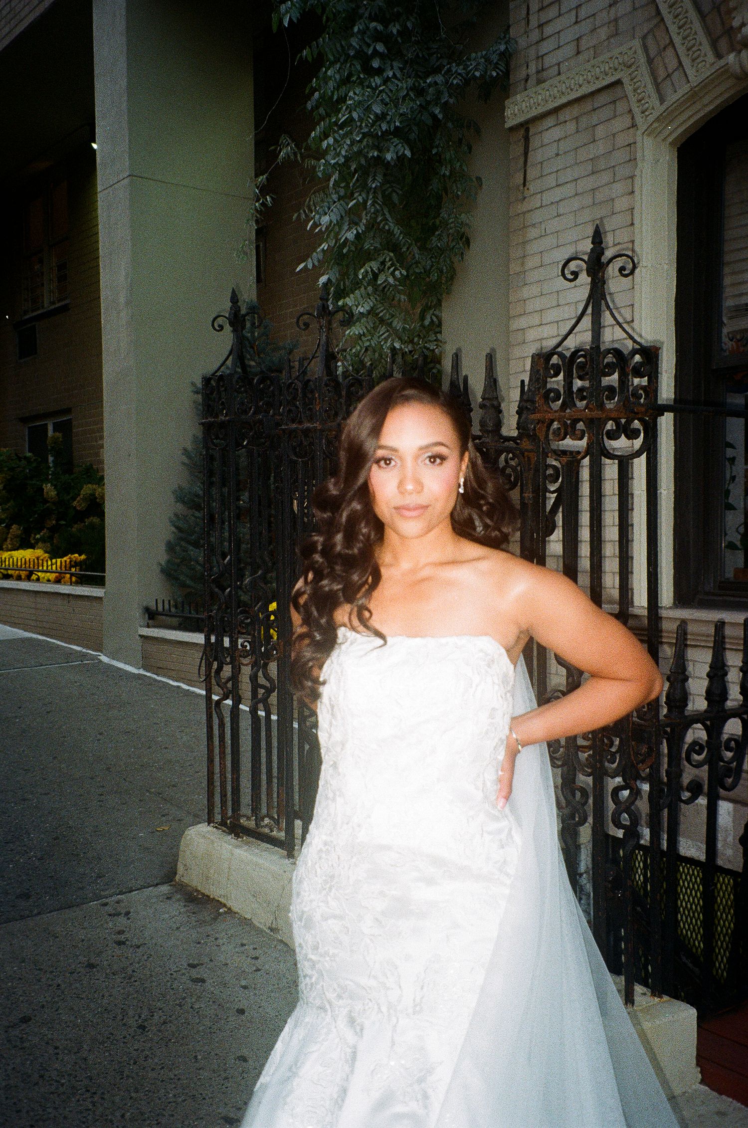 A bride in a strapless lace gown poses against an ornate iron gate and brick wall.