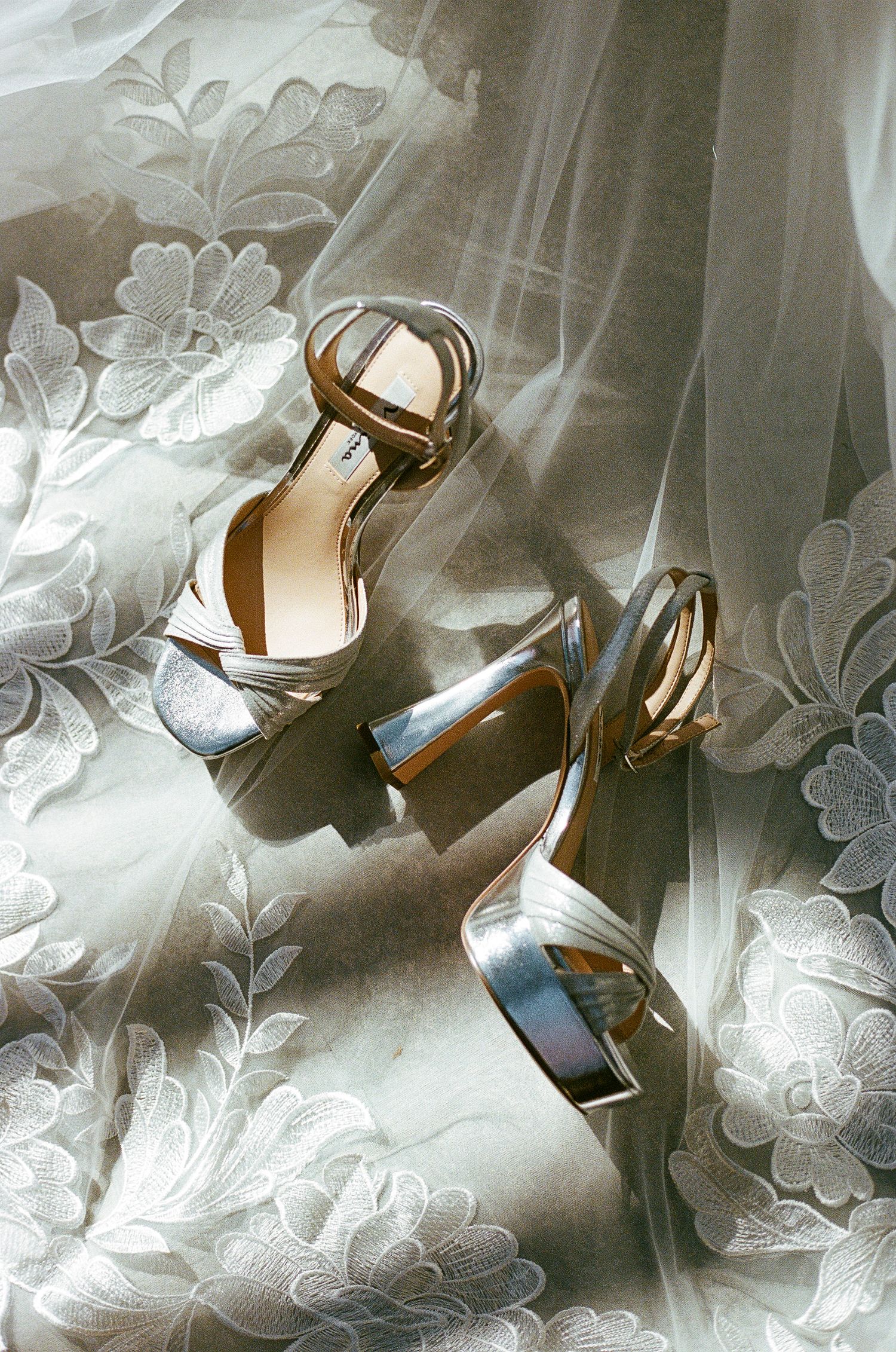 Silver bridal shoes photographed on white lace fabric with floral patterns.