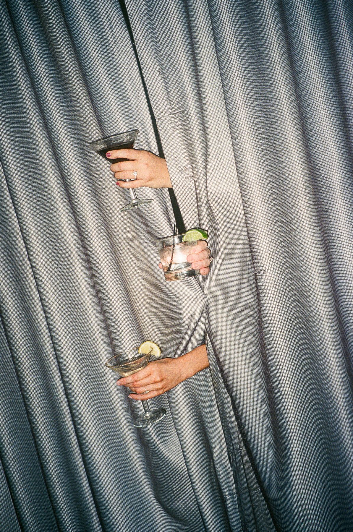 Three hands emerging from grey curtains holding cocktail glasses with lime garnishes at a party.