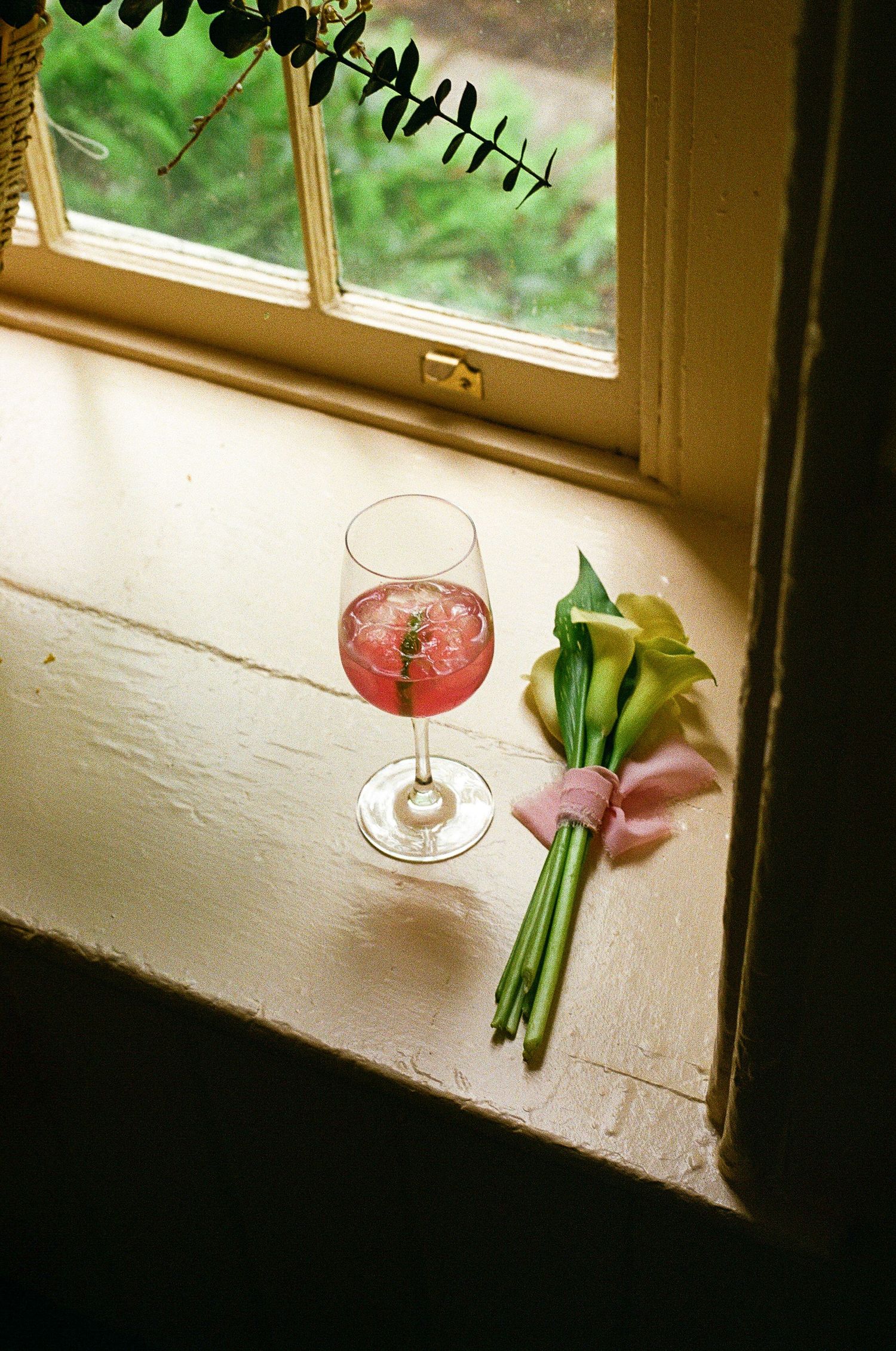 Pink cocktail in wine glass beside a small flower bouquet with ribbon on a sunlit windowsill.