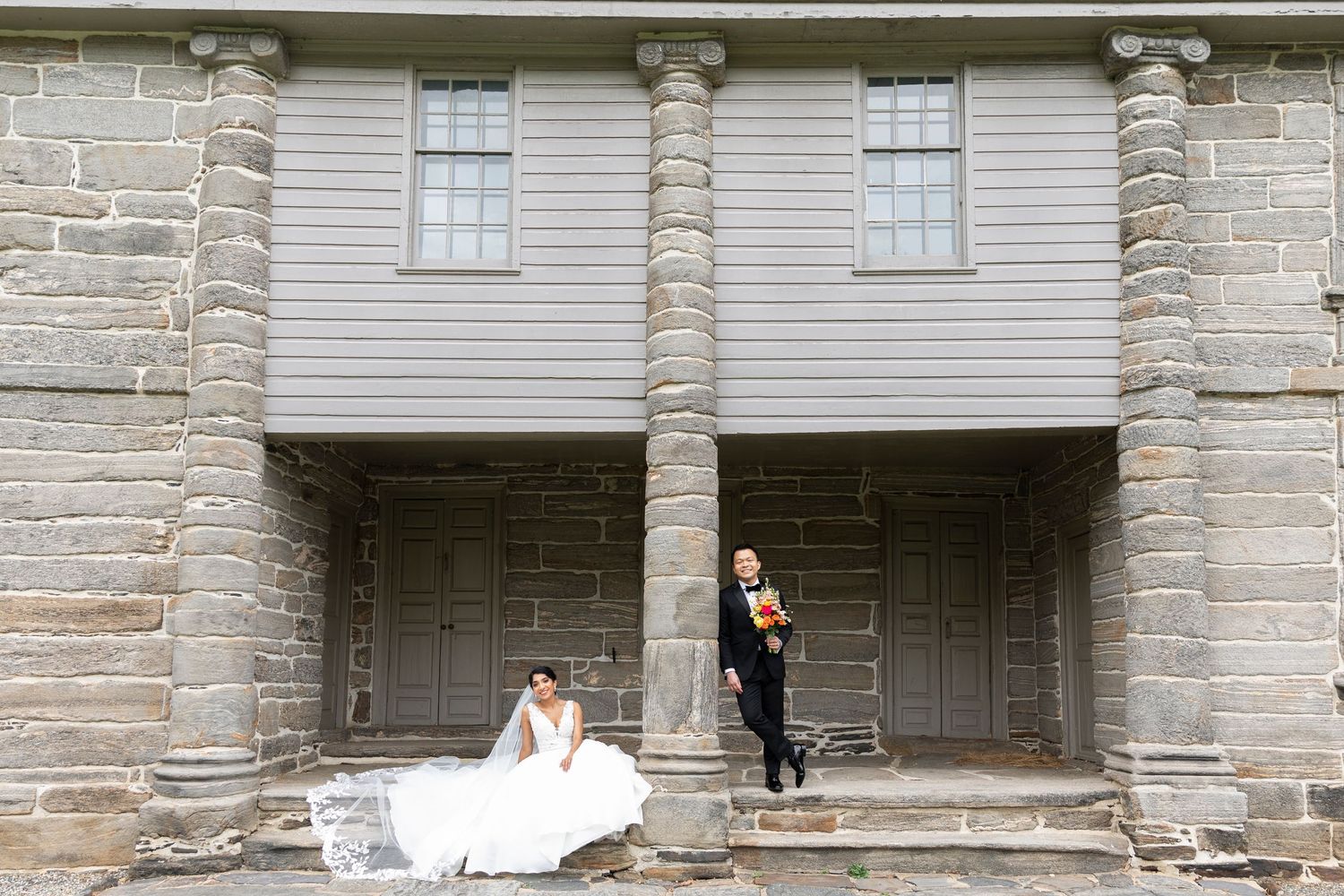 A wedding couple poses dramatically against a stone building with columns and dark doorways.