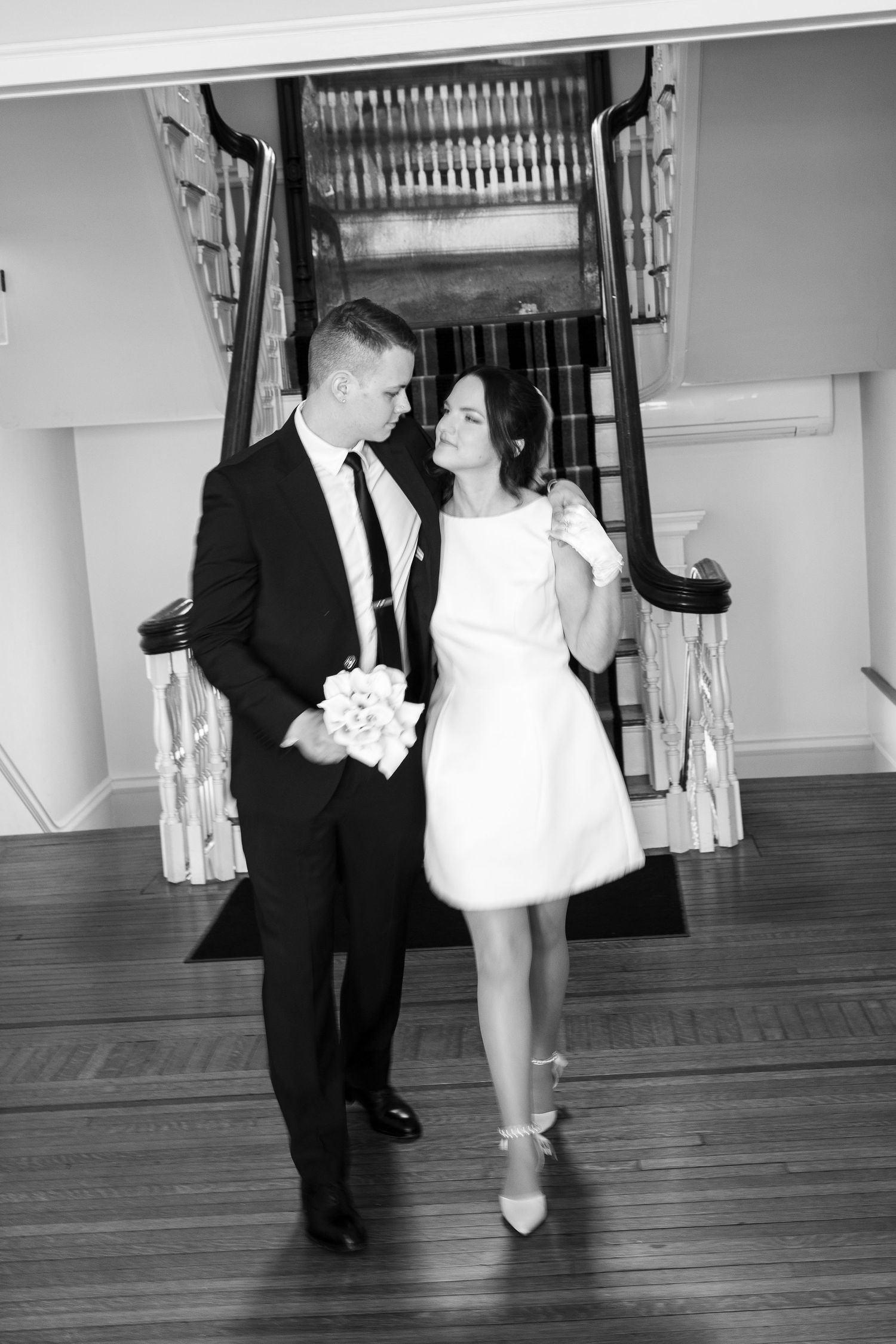 A romantic black and white photo of a couple in formal attire walking together on a hardwood floor.