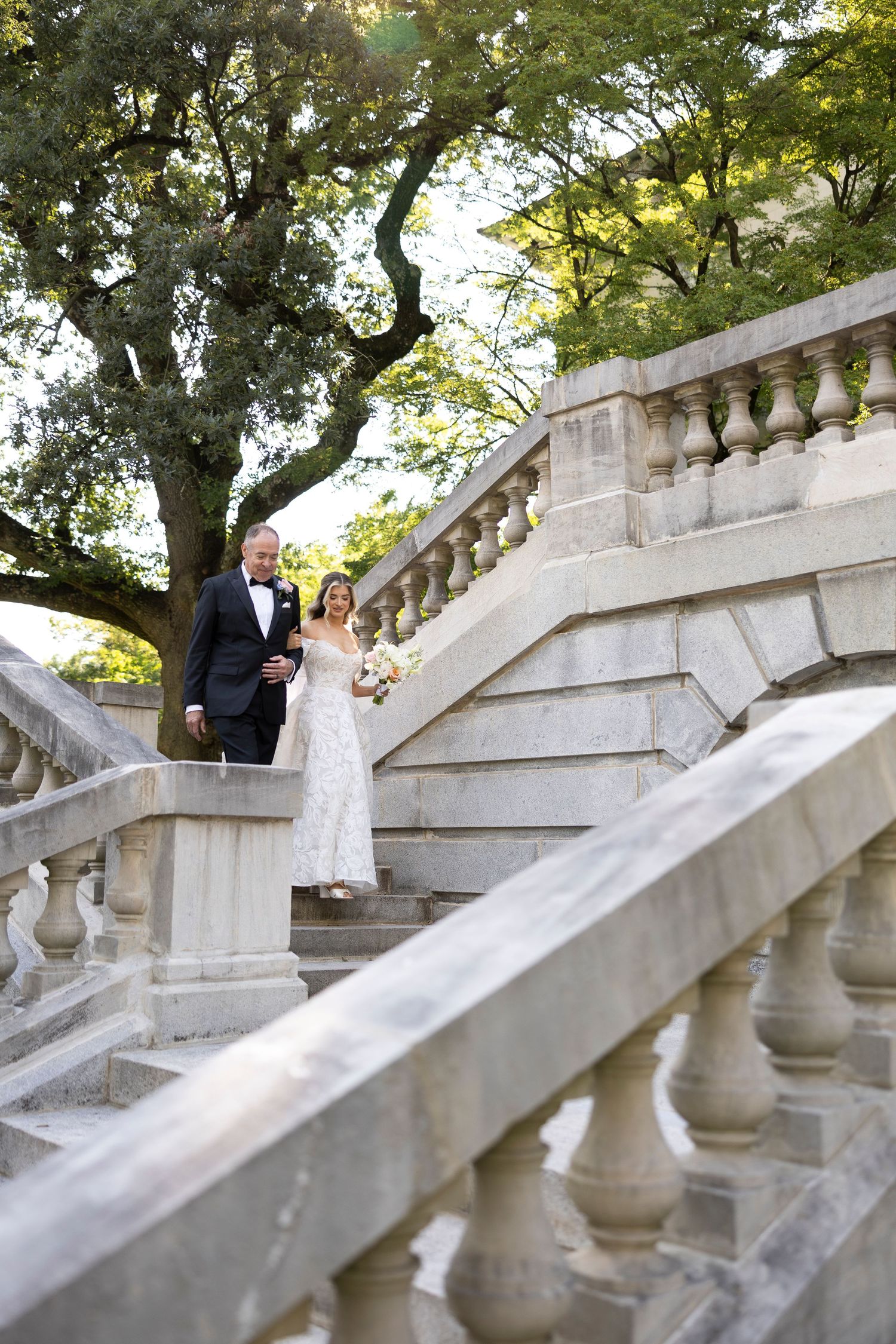 A couple descends an elegant stone staircase with ornate balusters surrounded by lush greenery.