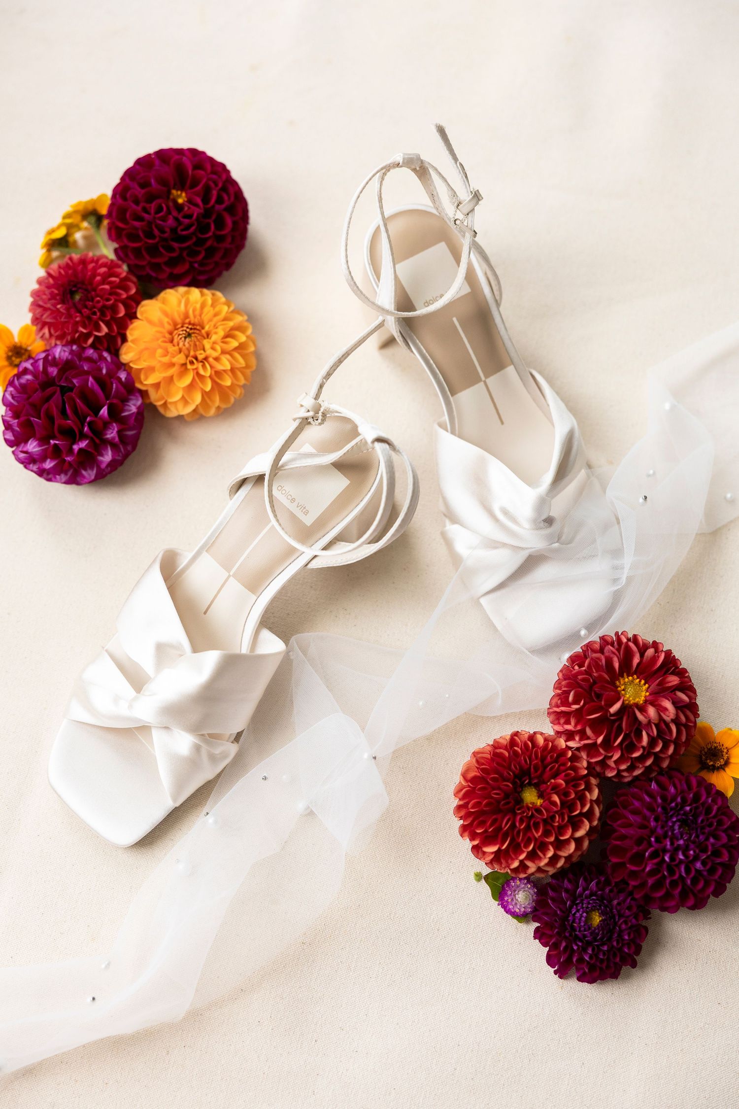 White bridal shoes arranged with deep burgundy and orange dahlias on a light background.
