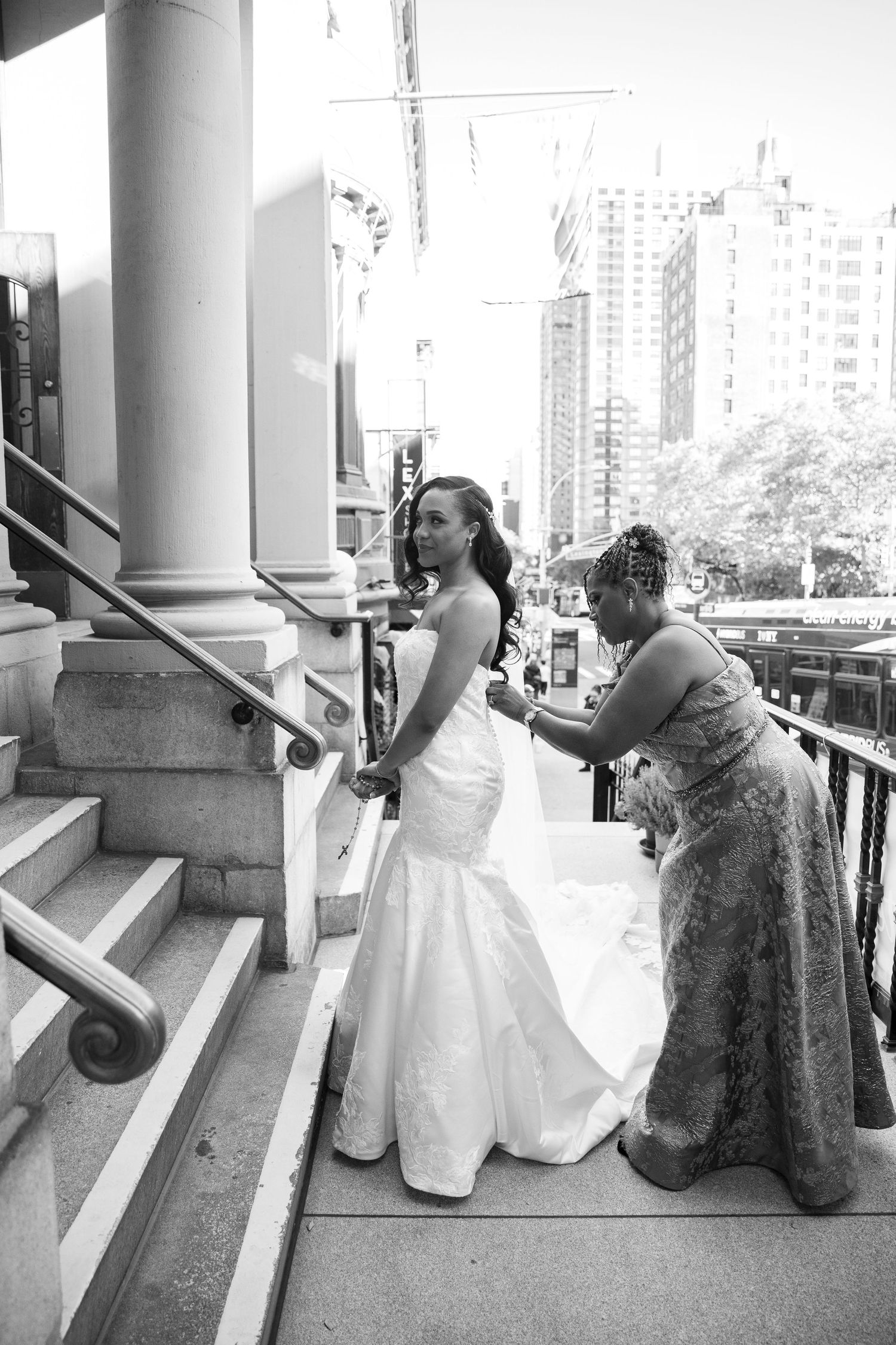 Wedding party posing for photos on outdoor steps with city buildings visible in the background.