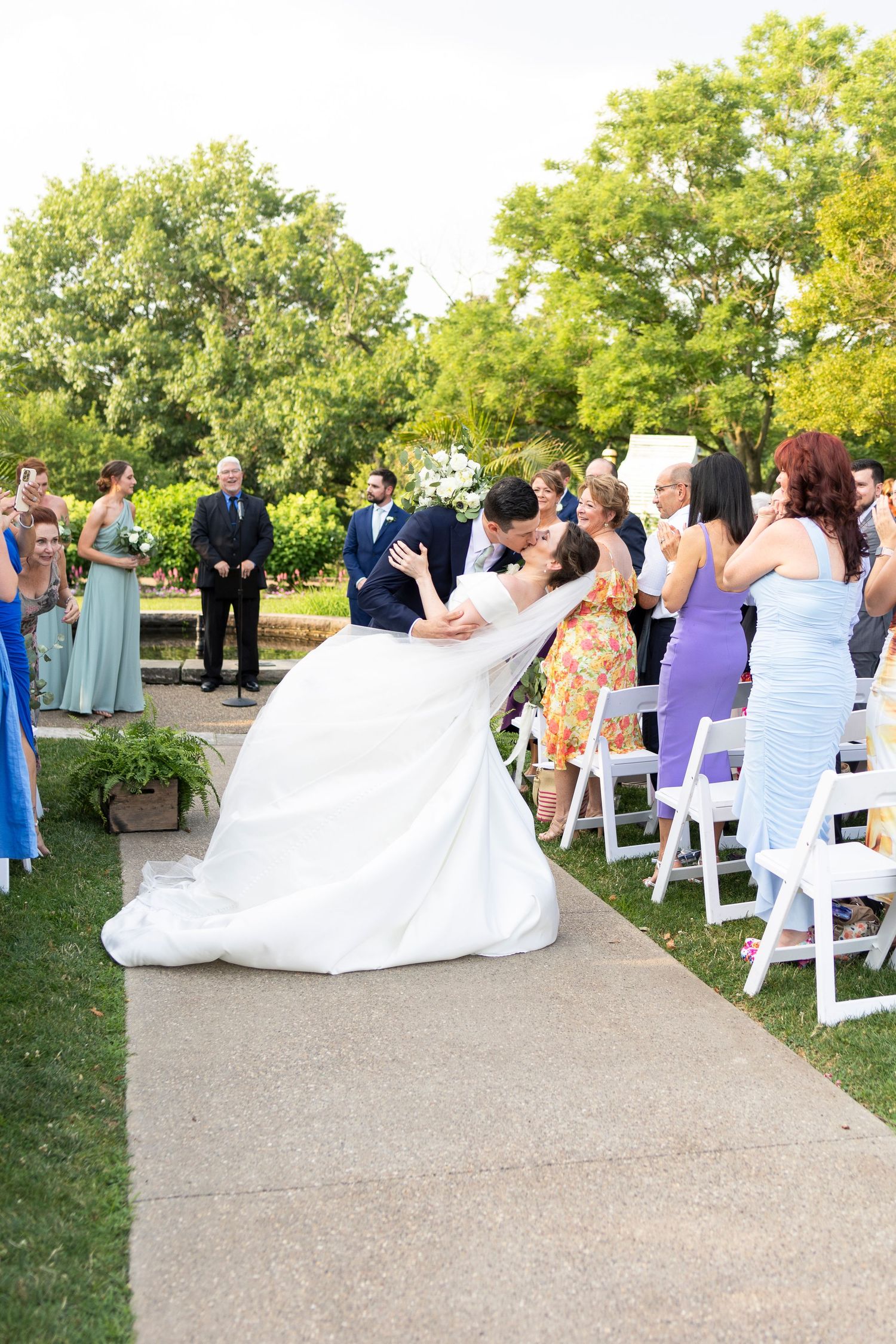 Outdoor wedding ceremony setup with white chairs and guests on a paved garden pathway.