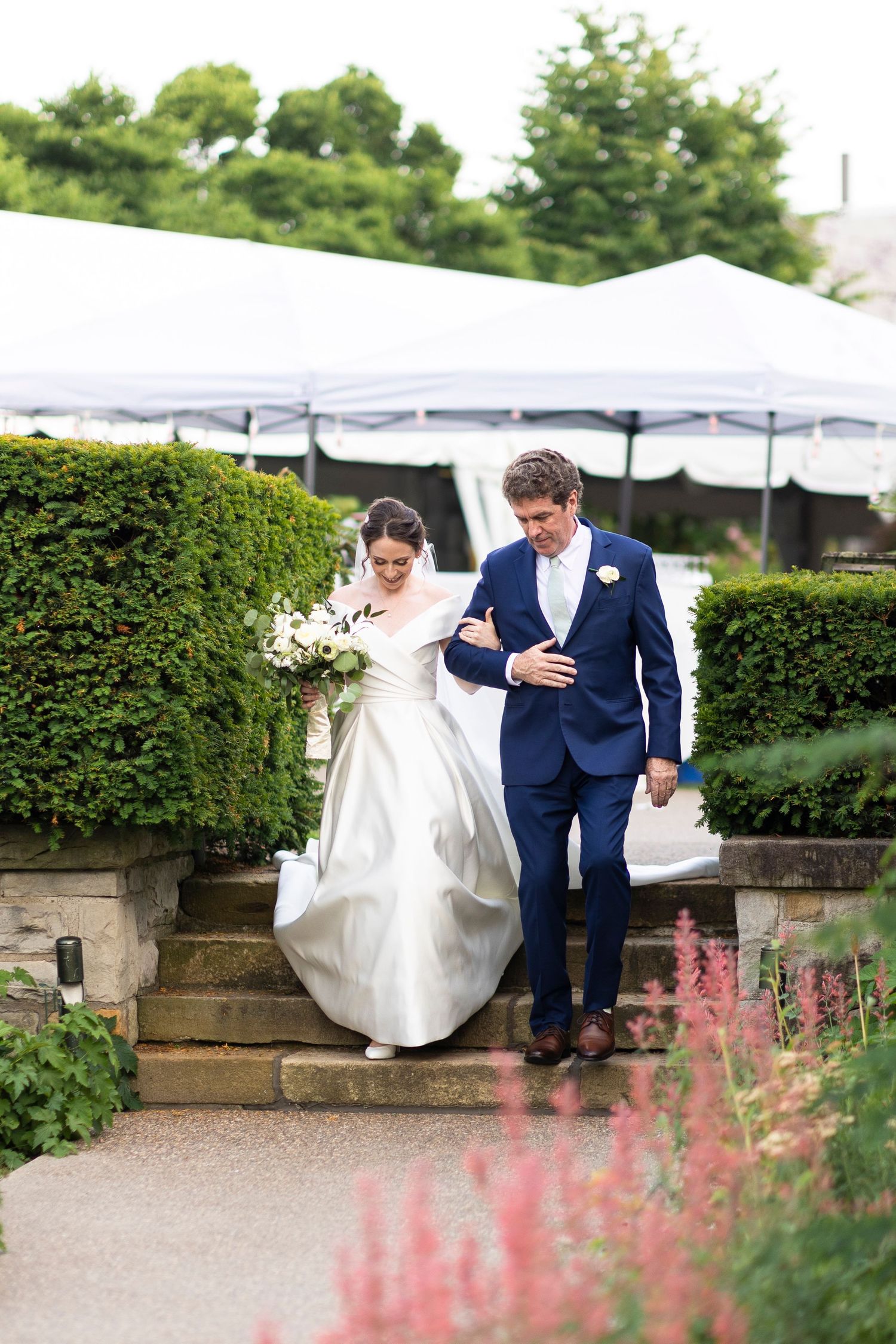 A couple walks along a garden path with pink flowers and hedges in the background.