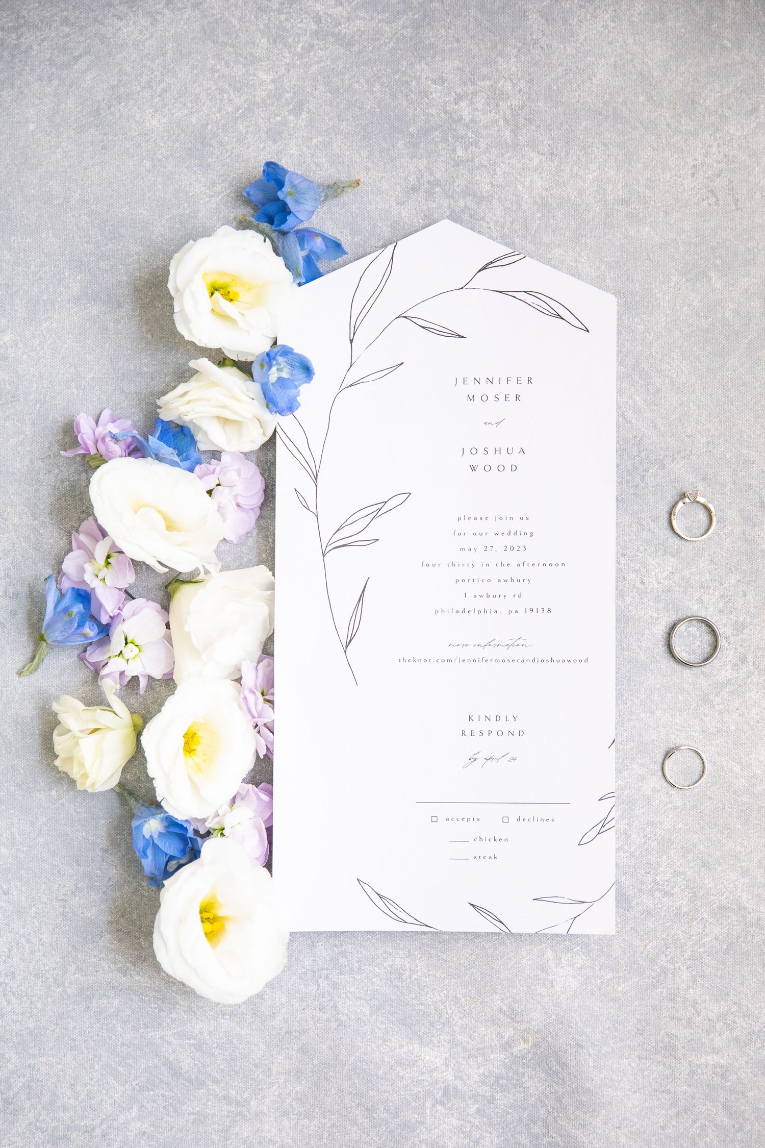 Minimalist wedding invitations with floral accents and wedding rings styled on a gray background.