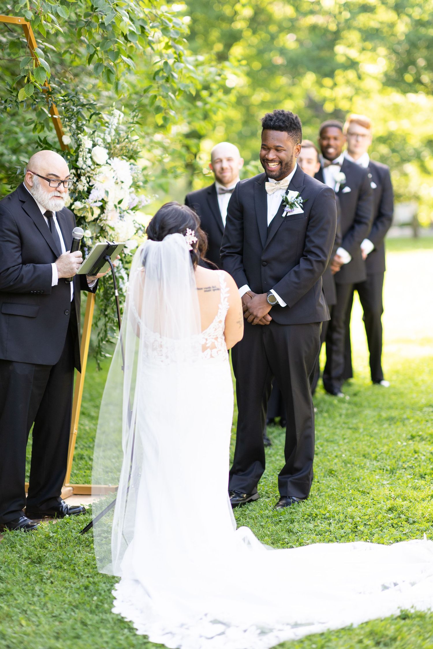 Wedding ceremony participants stand beneath a wooden arch decorated with white flowers.
