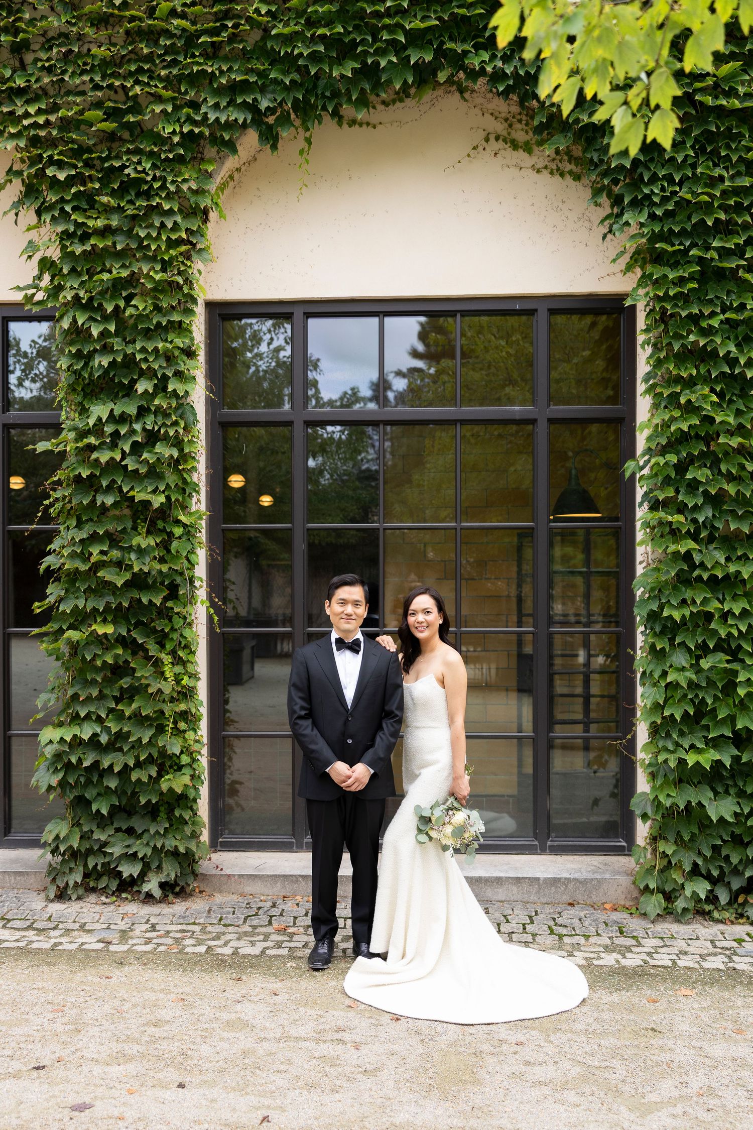 Wedding couple poses against ivy-covered building with large windows.