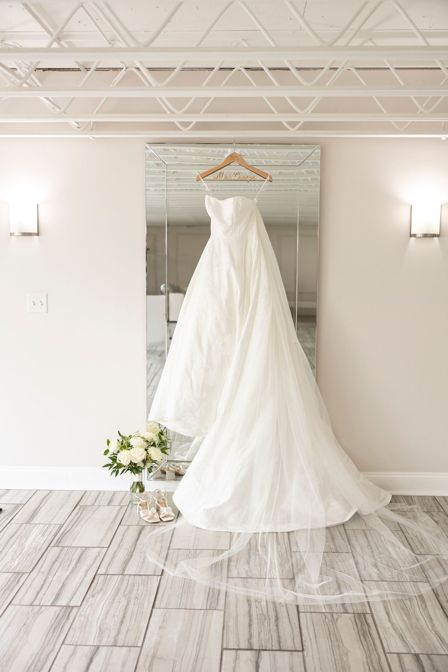 A white wedding dress hangs elegantly on display in a bright studio space with wood flooring.