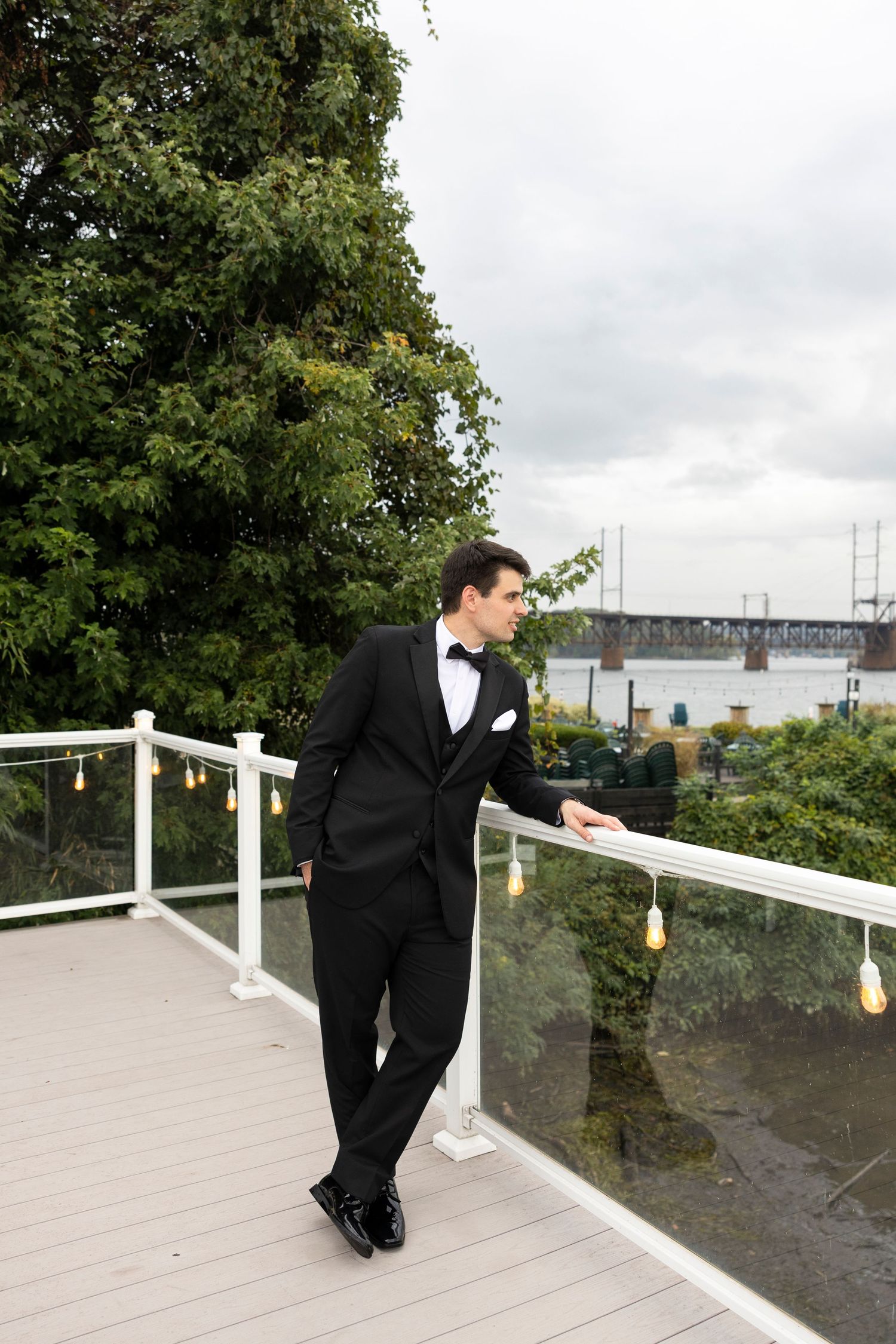 A person in formal attire walks along a white-railed deck with trees and a bridge in the distance.