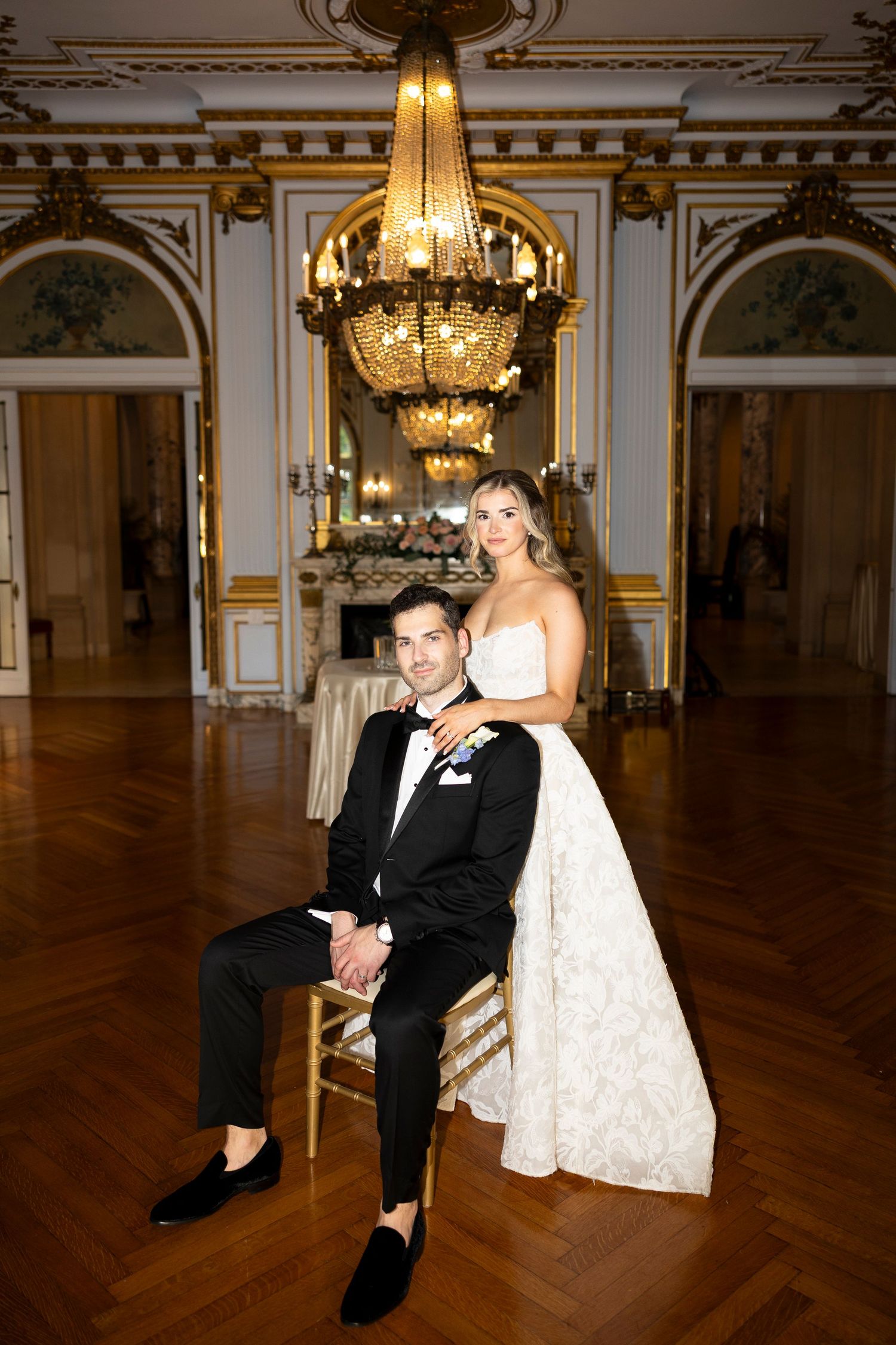 Elegant couple poses in a luxurious ballroom with ornate gold details and chandeliers.