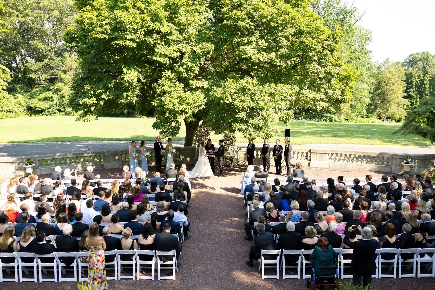Wide view of outdoor wedding ceremony with rows of white chairs and gathered guests.