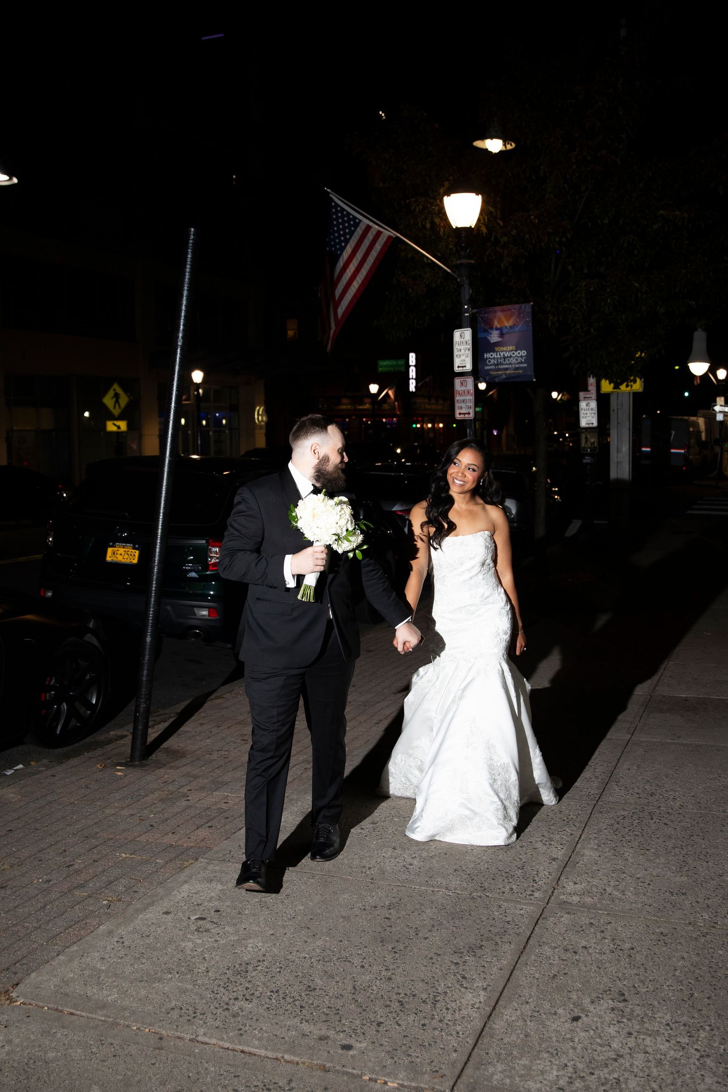 Wedding couple walking along a dimly lit sidewalk at night outside a venue.