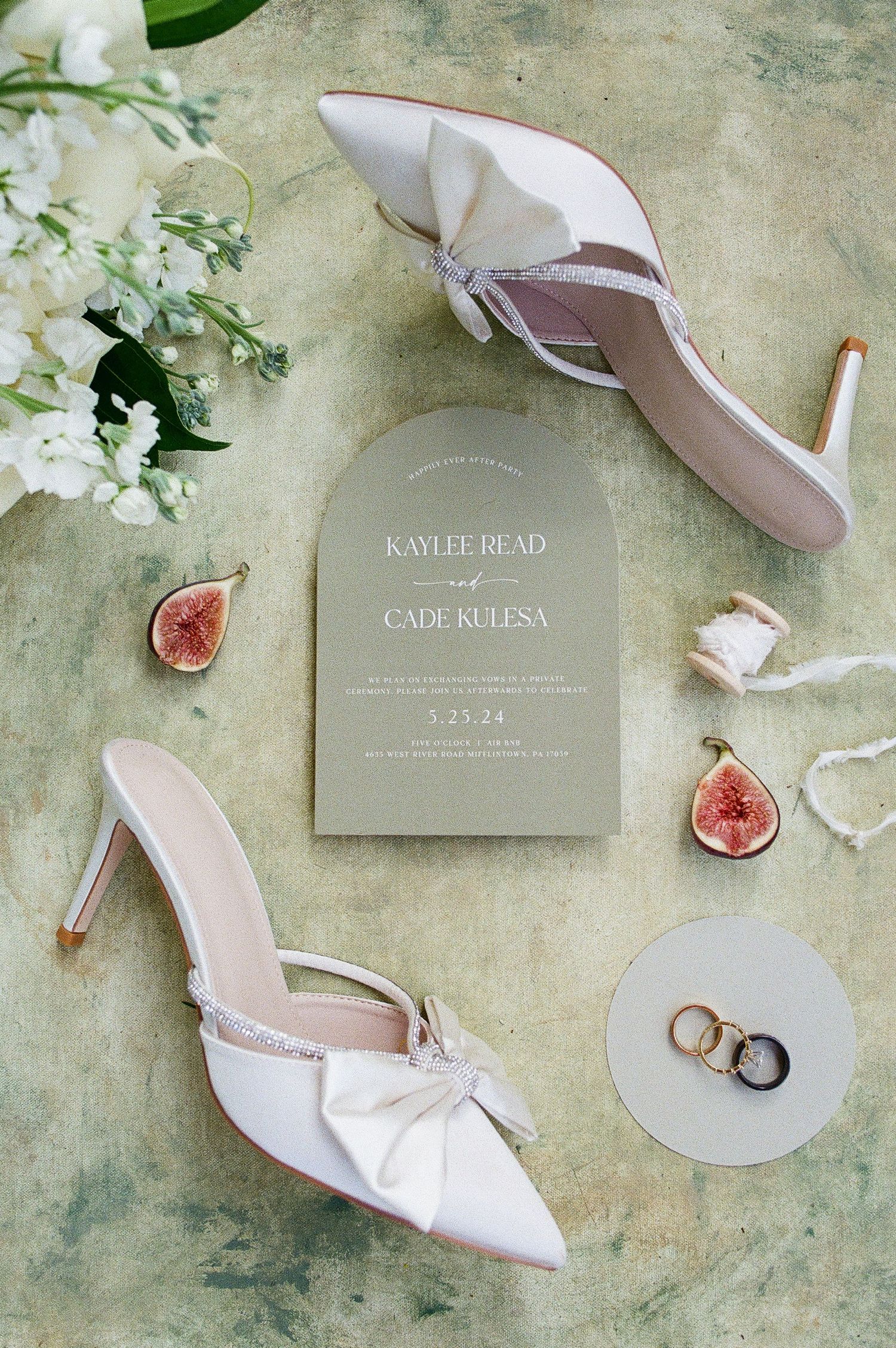 Wedding details flatlay with white shoes, rings, invitation and fresh figs on sage green background.