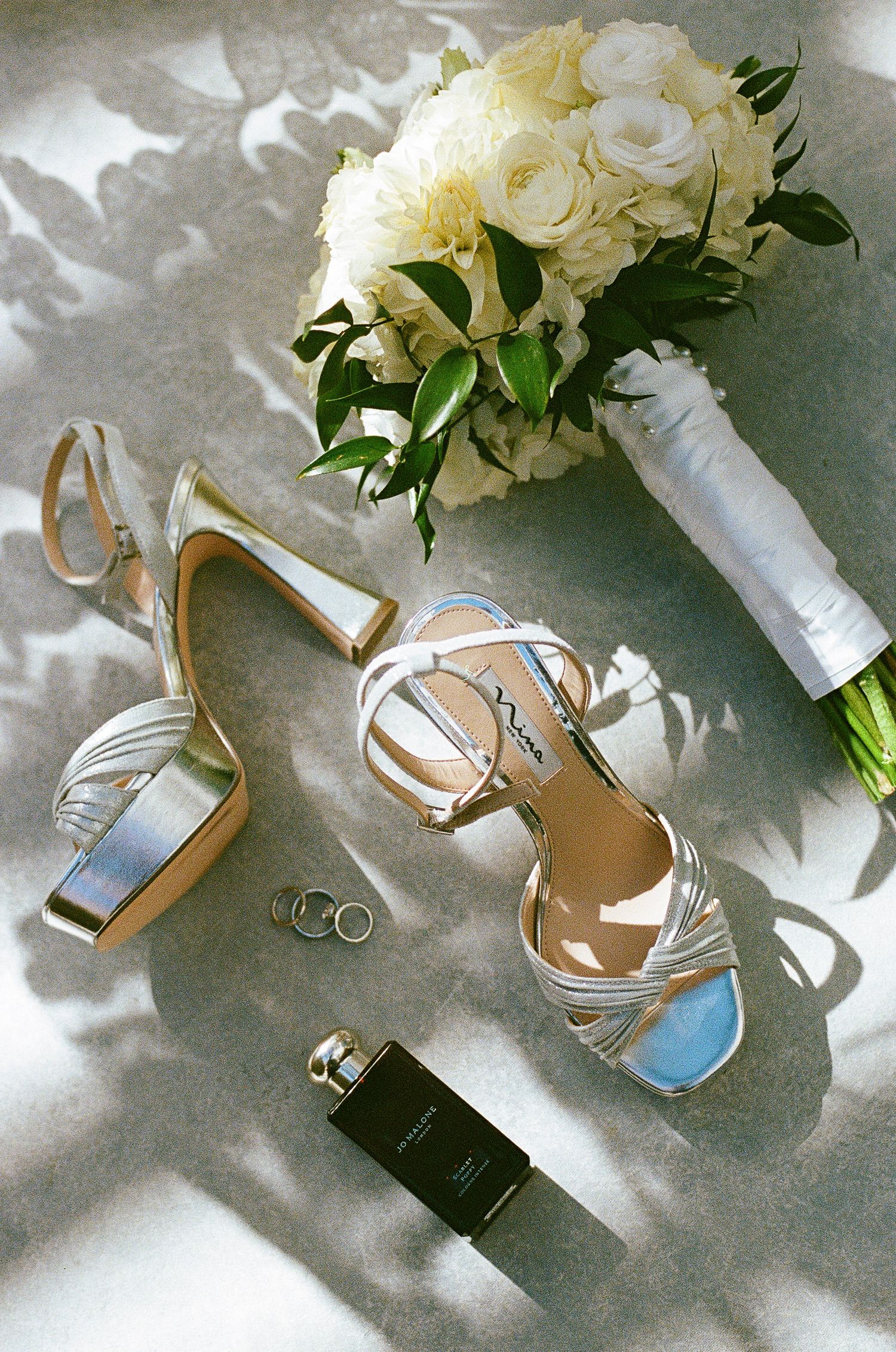 Wedding details including white rose bouquet, rings, and silver heels arranged artistically.