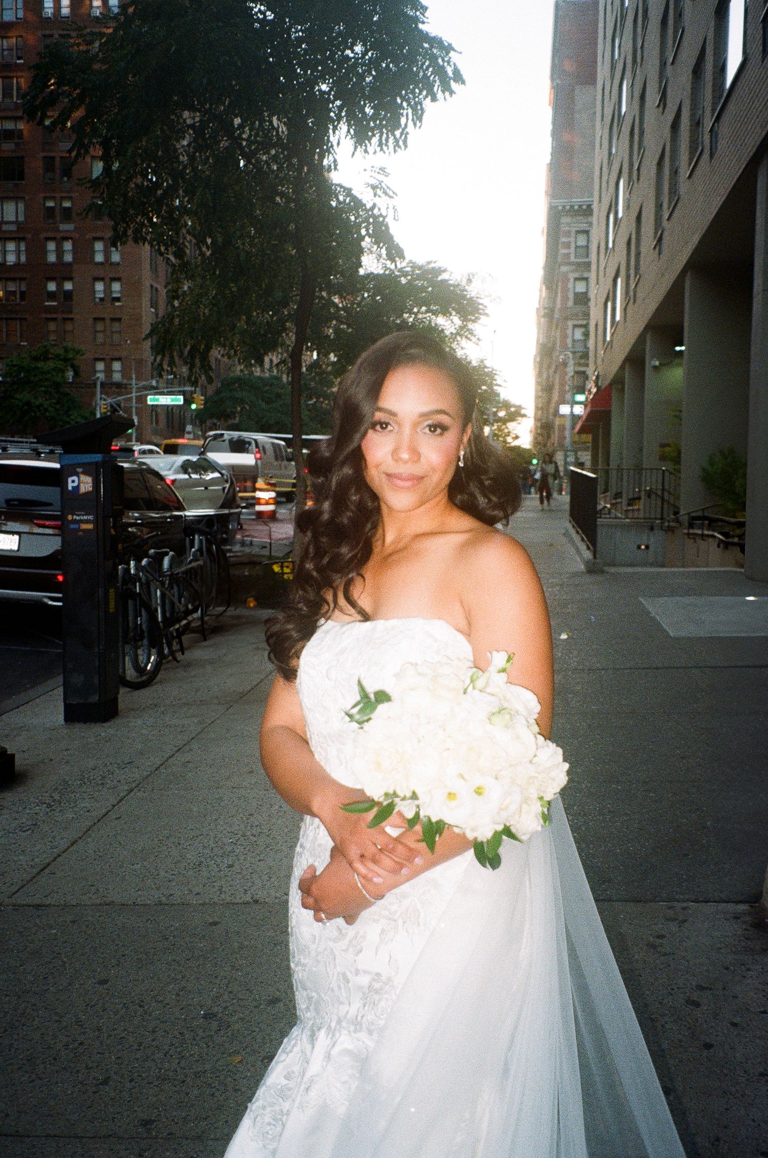 A bride in a white wedding dress holding a bouquet poses on a city street at dusk.